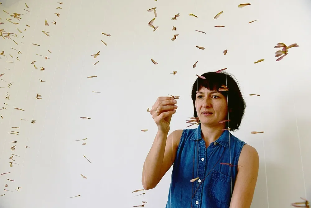 A woman in a sleeveless denim shirt is looking at small, hanging flower buds or pods suspended from thin strings against a plain white background.