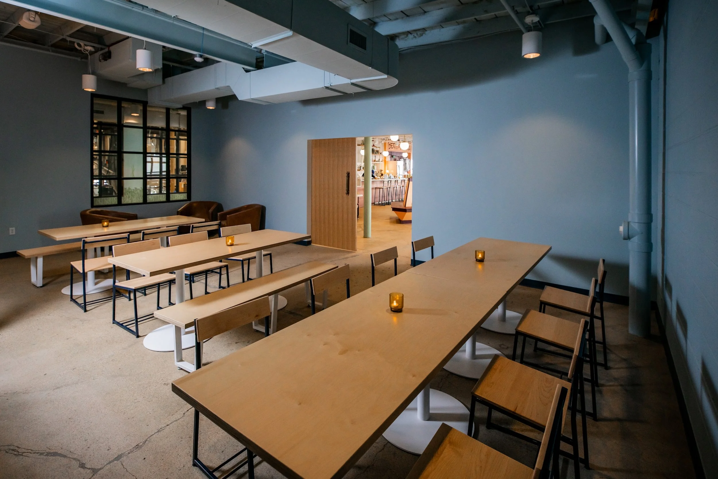 Inside Honest Friend Brewing's modern, minimalistic sitting area with wooden tables, chairs, and small candlelights, adjacent to a bar visible through a door, with exposed ceiling pipes and soft lighting.