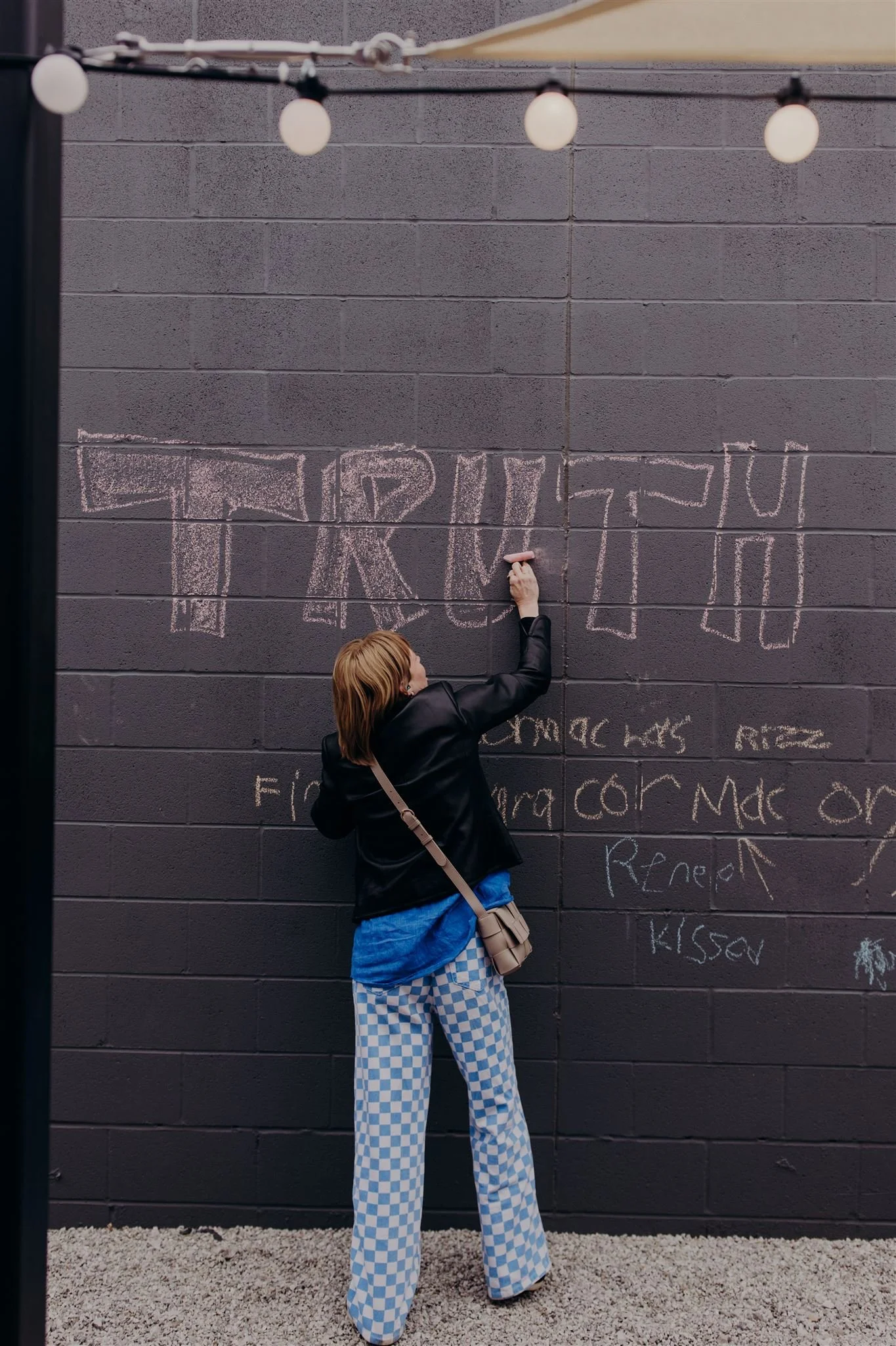 Person writing on a black wall with pink chalk, with large checkered blue and white pants, a black jacket, and a small crossbody bag. String lights hang above.