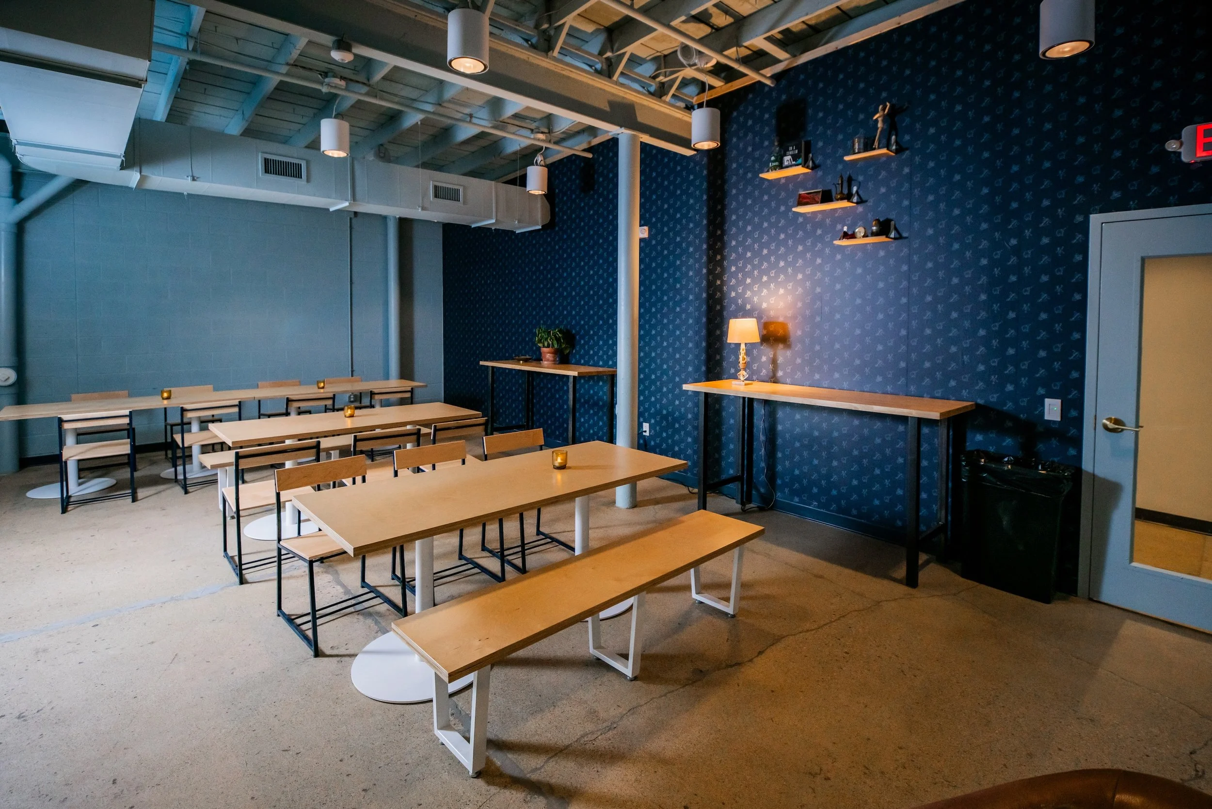 Inside Honest Friend Brewing's seating area with wooden tables, benches, and chairs, dark illustrated patterned wall with wall art and shelves, table lamp, and candle holders on tables.