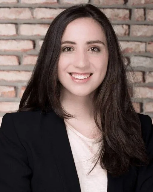A young woman with long dark hair smiling, wearing a black blazer and light-colored top, standing in front of a brick wall.