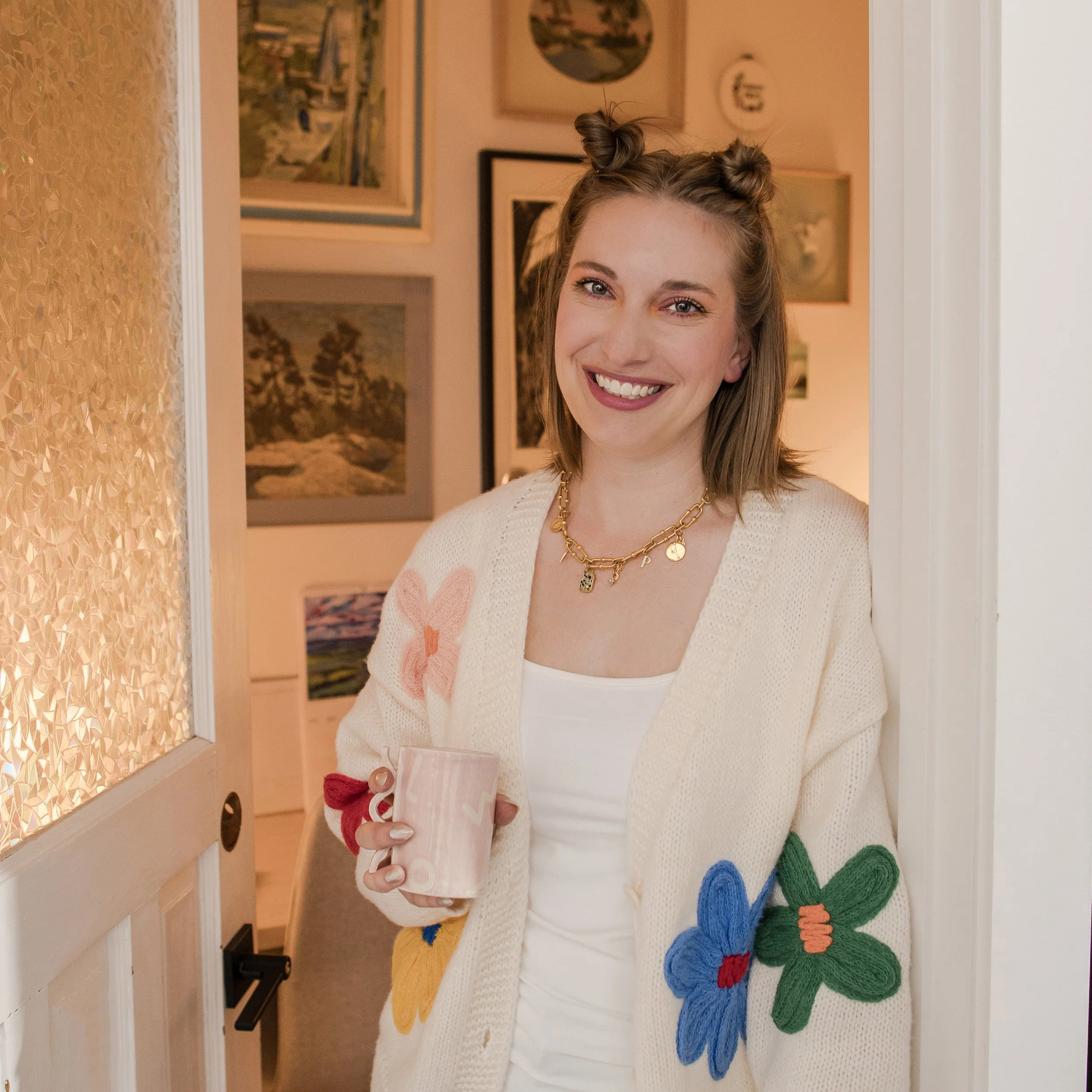 Headshot of Nicole Winski, a woman with shoulder-length medium blonde hair, wearing a cream sweater and smiling softly against a door frame to an office with artwork on the wall