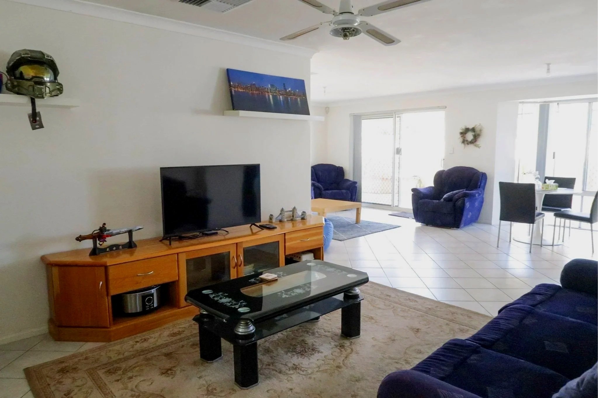 Living room with a dark blue couch, armchair, and a dining table with chairs. There is a TV on a wooden stand, a ceiling fan, sliding glass doors, and a wall decoration. A shelf holds a helmet and model figures.