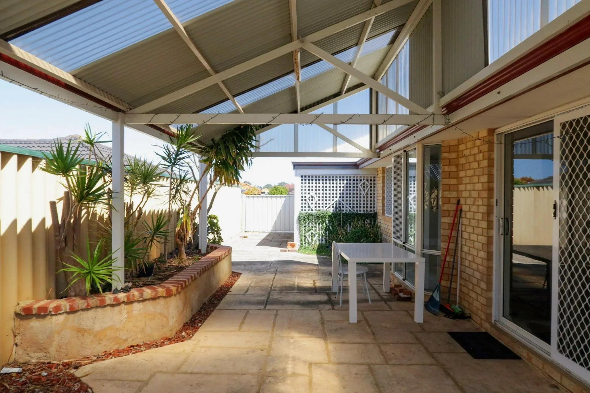 Backyard patio area with a covered roof, potted plants, a white table with chairs, brick border garden bed, and sliding glass doors.