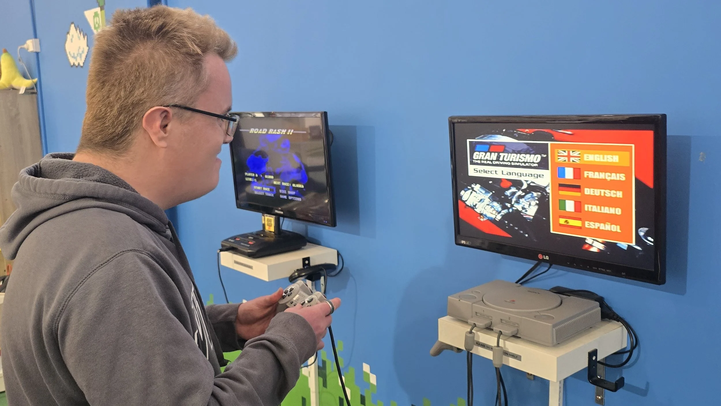 A young man with glasses and a hoodie is playing a racing video game on an arcade machine, holding a controller, with a blue wall and gaming screens in the background.
