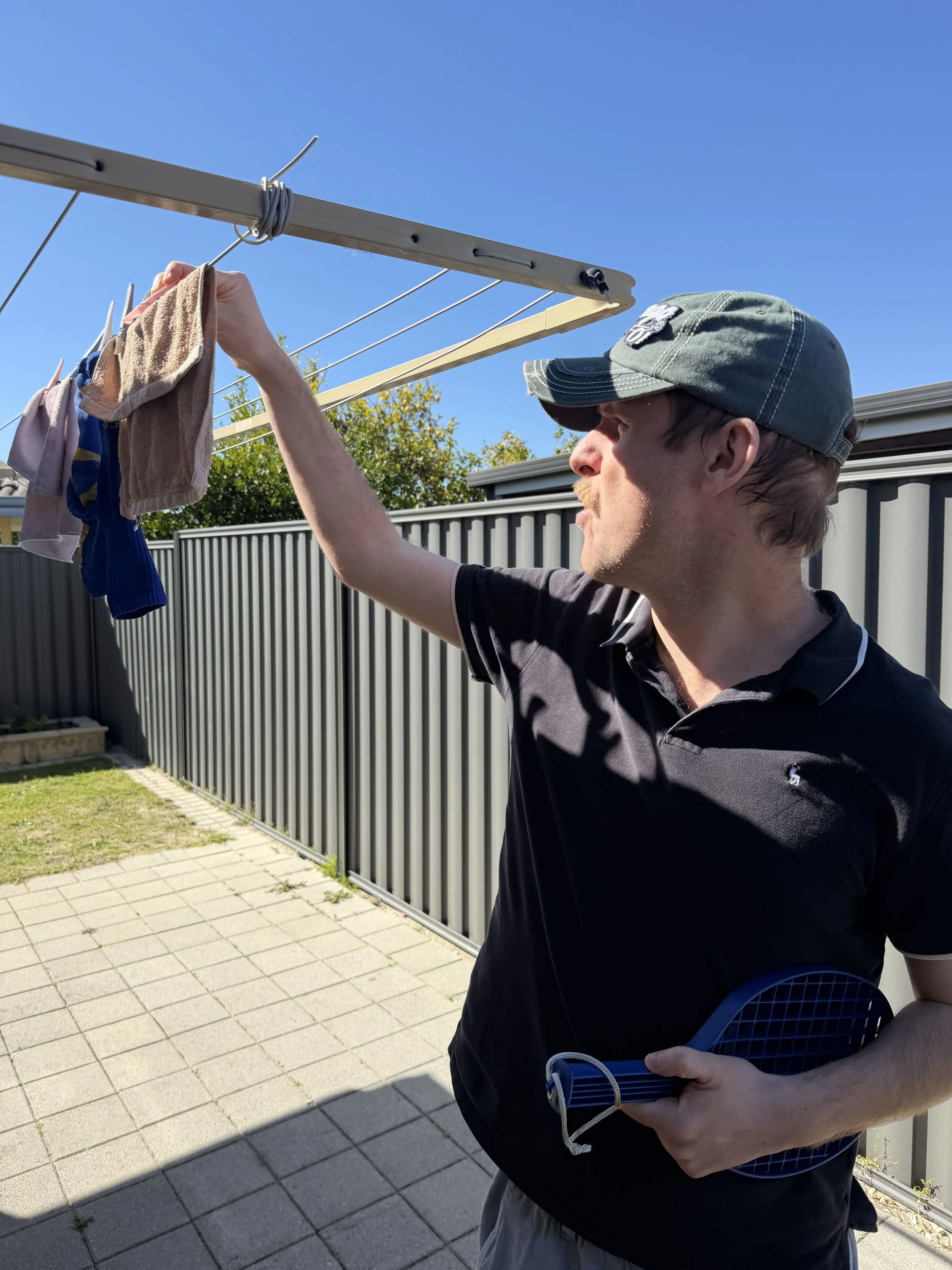 A man hanging clothes on a clothesline outdoors with a blue sky in the background.