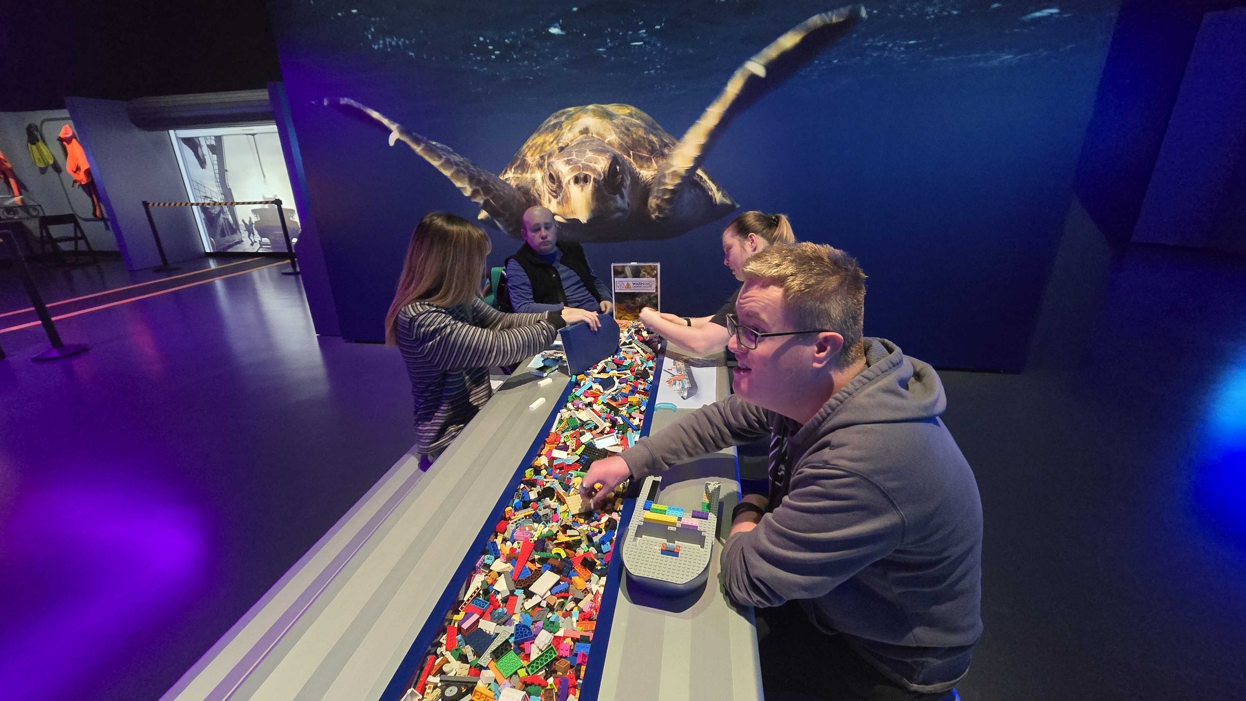 People playing with LEGO bricks on a table in a dark exhibit with a large projected image of a sea turtle on the wall behind them.
