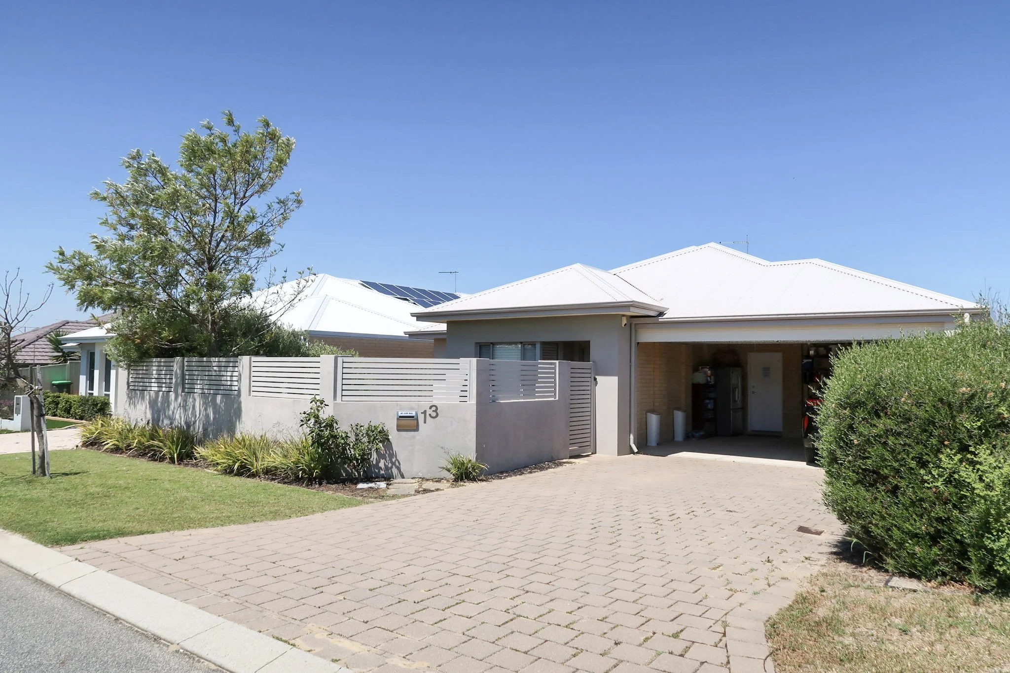 Modern house with solar panels on the white roof, surrounded by trees and bushes, with a brick driveway and a small front yard.