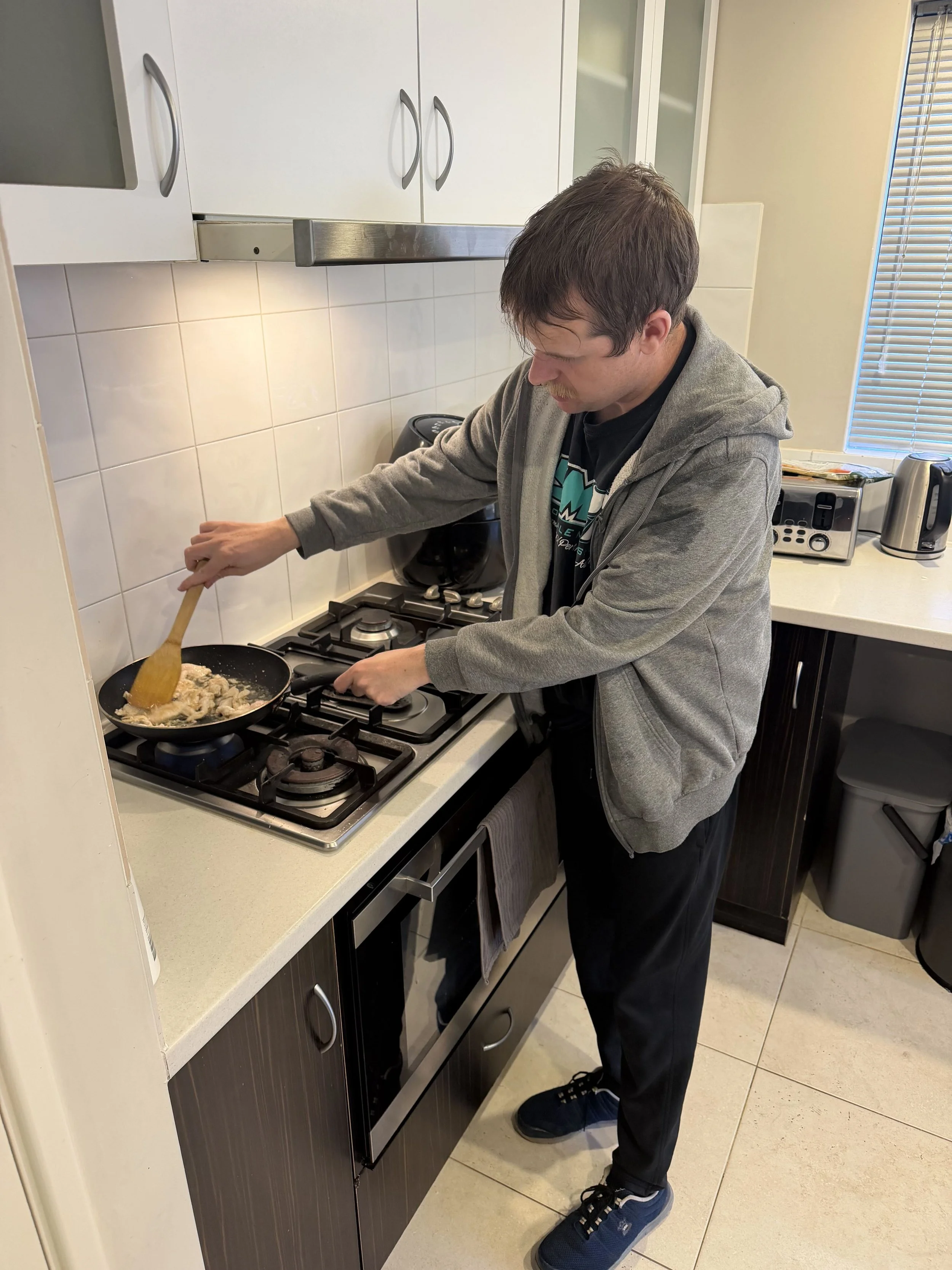 A man cooking at a stove in a kitchen, wearing a gray hoodie, black pants, and blue sneakers, with kitchen appliances and cabinets around him.