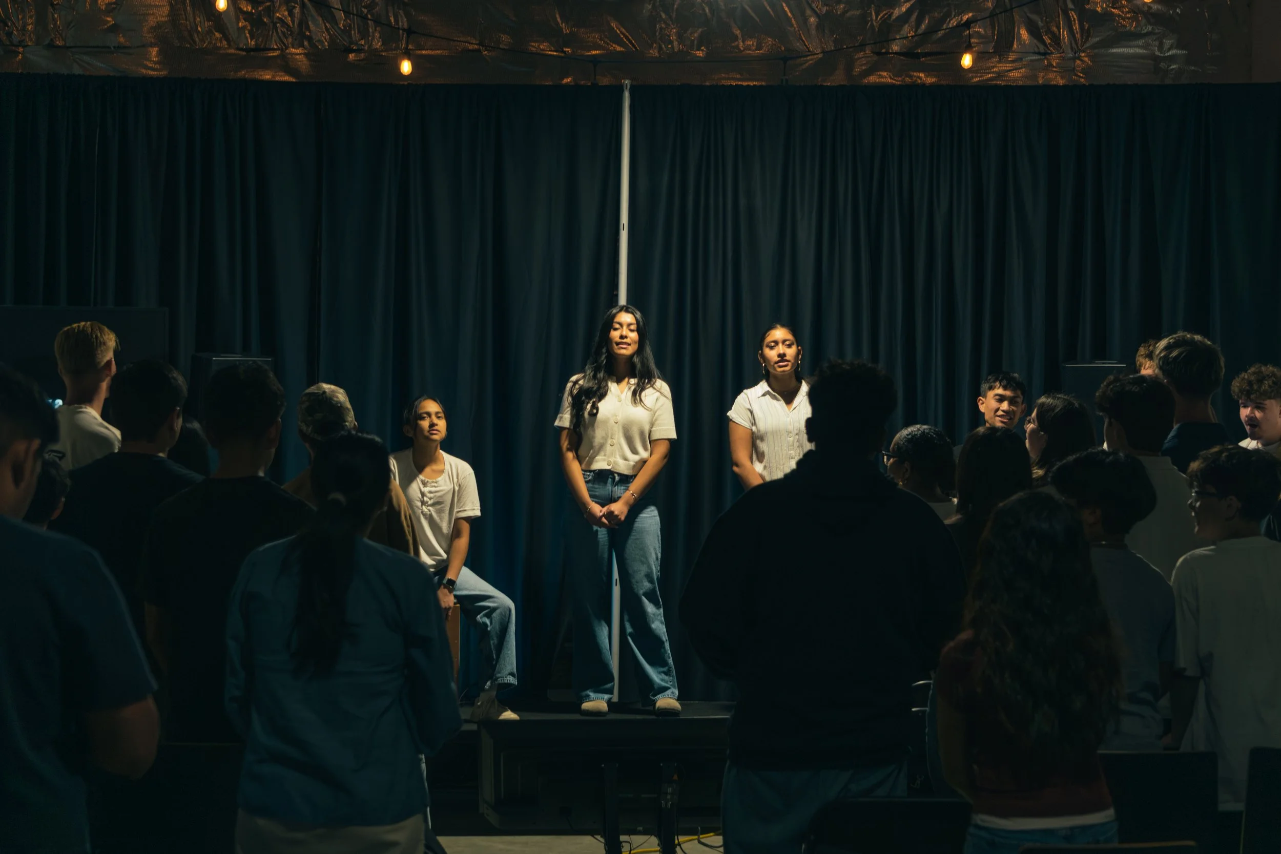 A group of young people on a stage with a dark blue curtain, some standing and one sitting, while an audience watches.