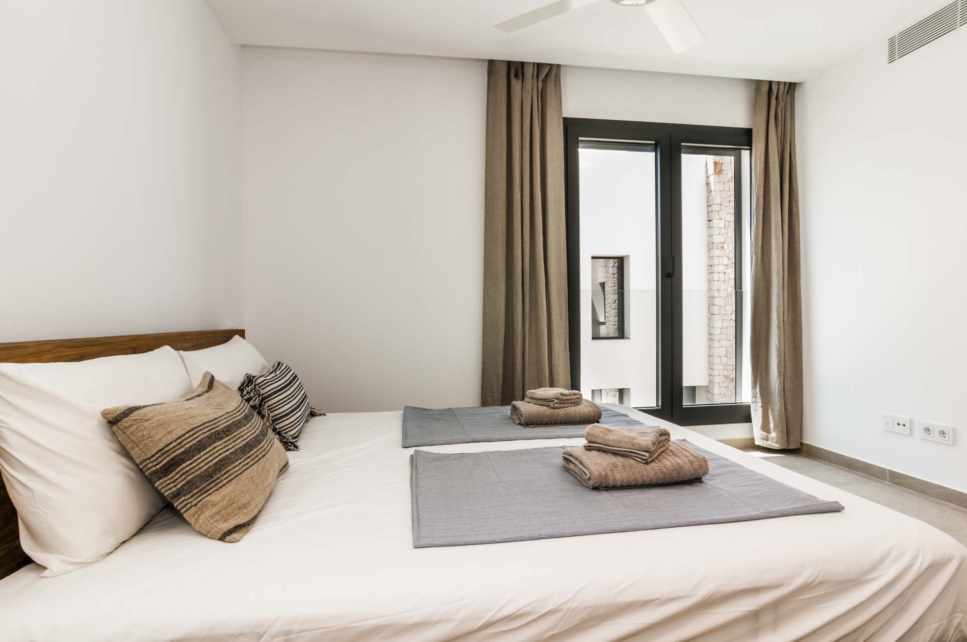 Modern bedroom with a double bed, striped pillows, folded towels on gray mats, large window with beige curtains, and a ceiling fan.