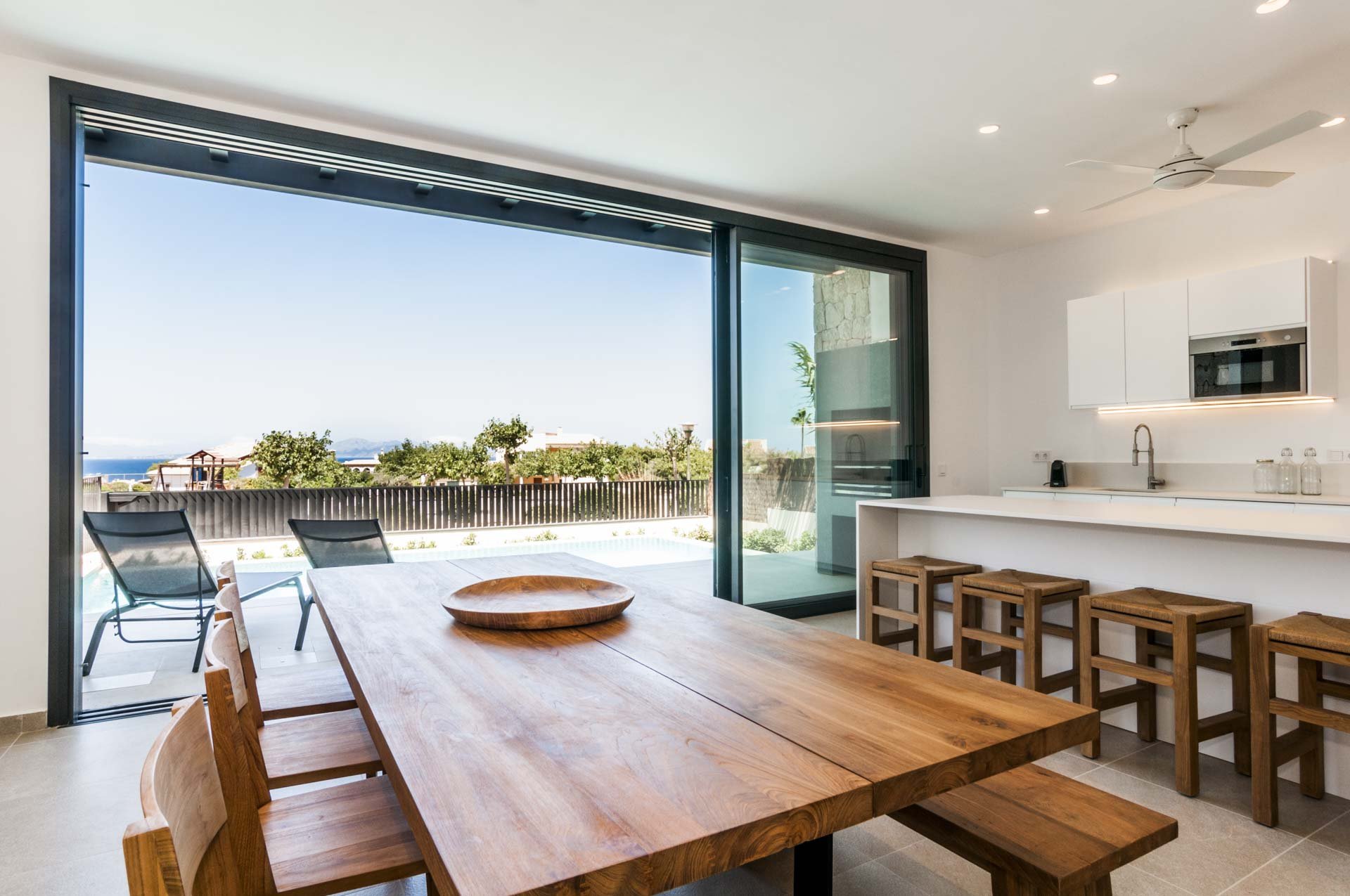 Modern kitchen and dining area with wooden table, stools, and large sliding glass doors opening to patio with pool and outdoor seating.