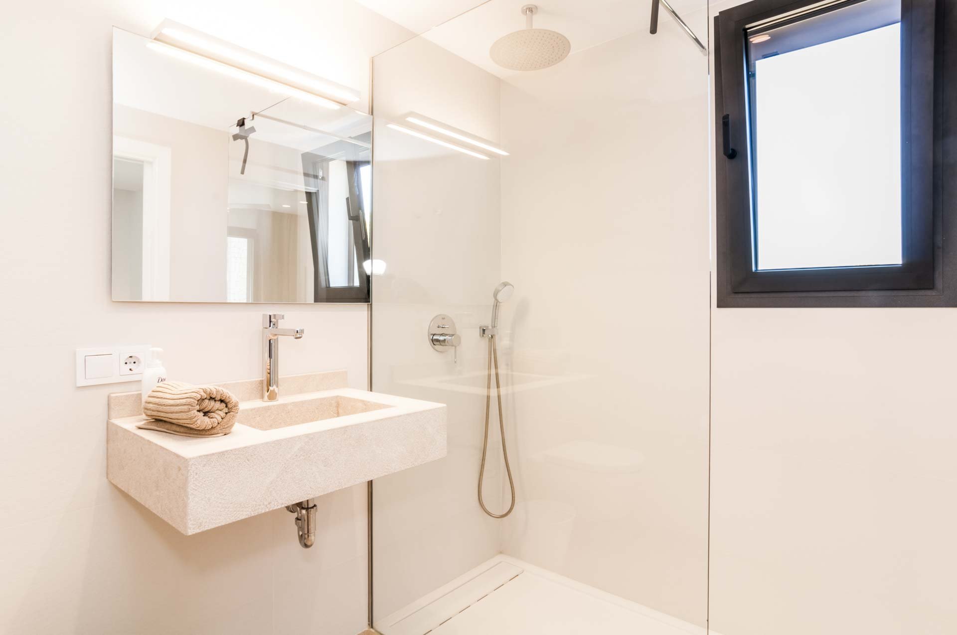Modern bathroom with a beige sink, wall-mounted faucet, showerhead, and a small window. A towel is folded on the sink with soap nearby. A large mirror is above the sink illuminated by overhead lighting.