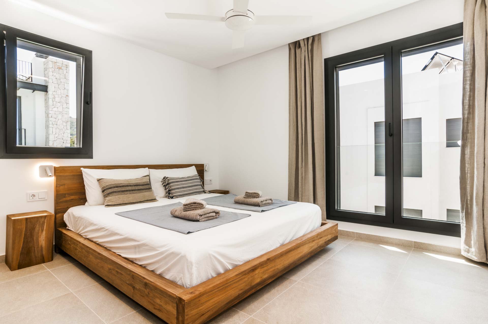 Modern bedroom with wooden bed frame, white bedding, striped pillows, two windows, beige curtains, and folded towels on the bed.