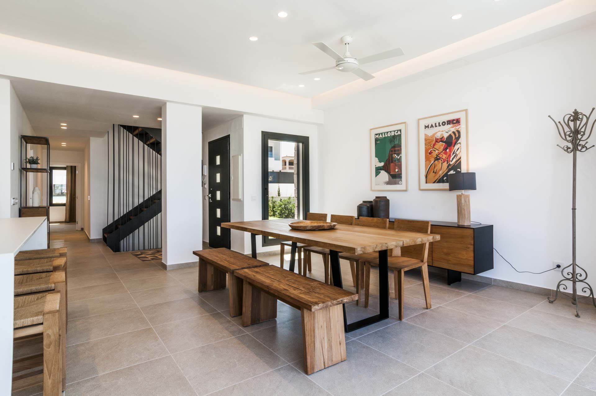 Modern dining room with wooden table, benches, and chairs, featuring minimalist decor, a ceiling fan, a staircase, and framed posters on the wall.