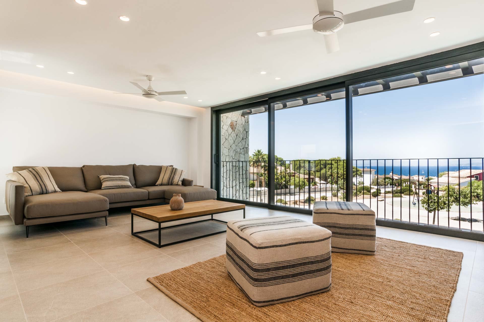 Modern living room with sectional sofa, two ottomans, coffee table, ceiling fans, and large windows showing an ocean view.