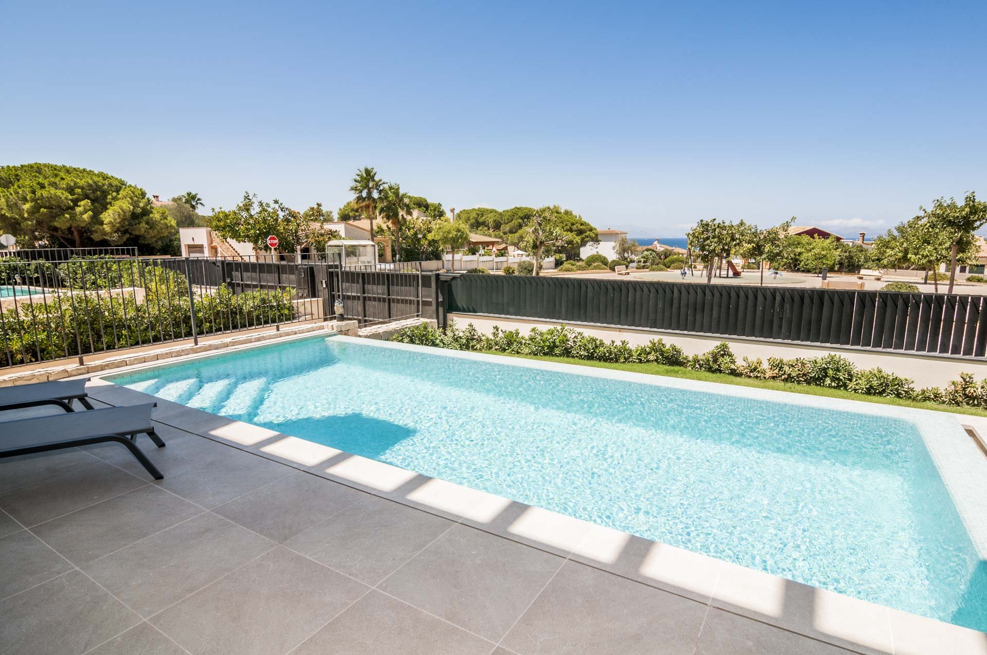Outdoor swimming pool with clear water, surrounded by a tiled patio, a black fence, and greenery; residential area in the background with trees and houses under a clear blue sky.
