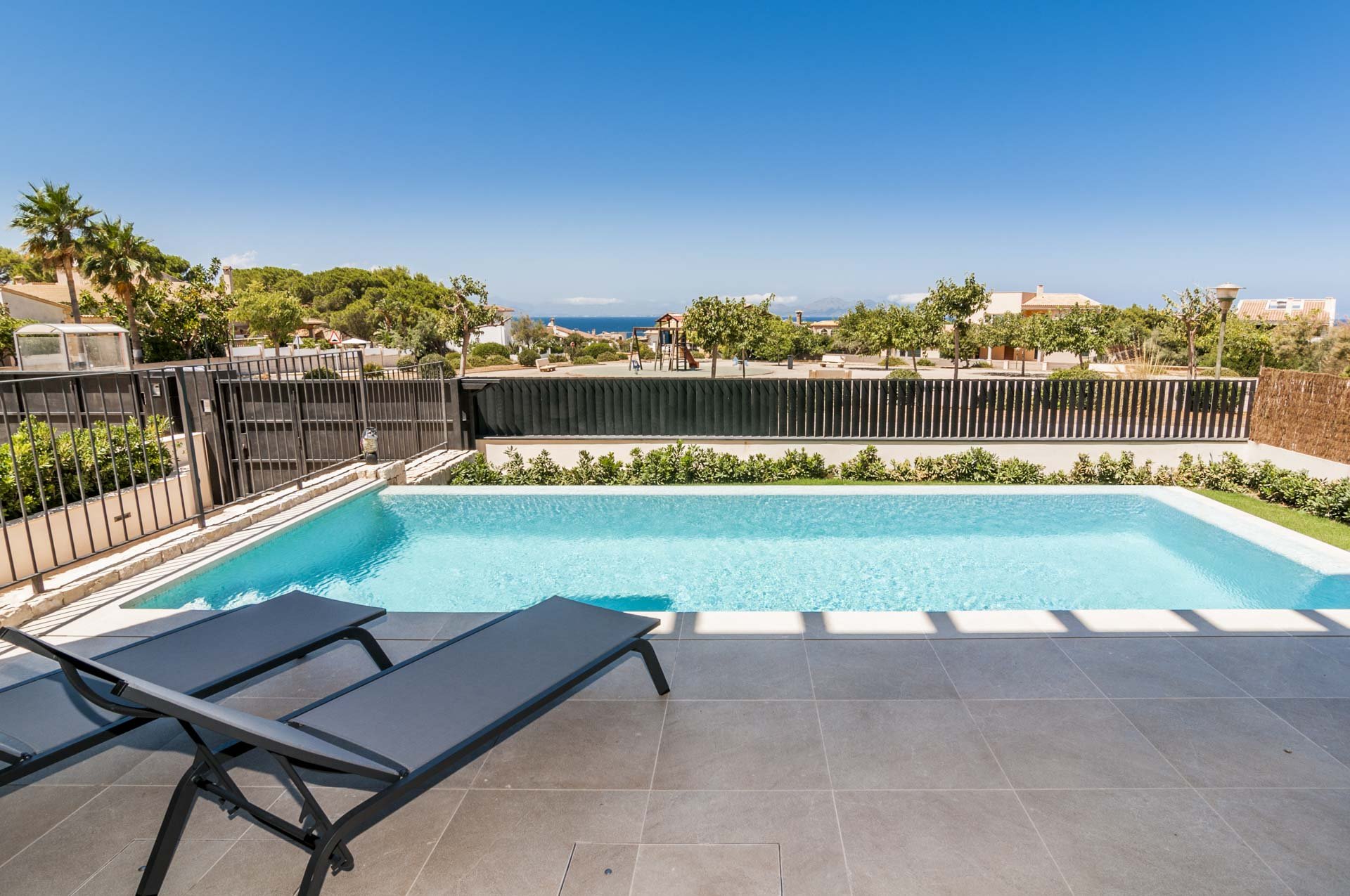 Backyard pool with sun loungers, surrounded by a fence, greenery, and a view of trees and houses in the distance.
