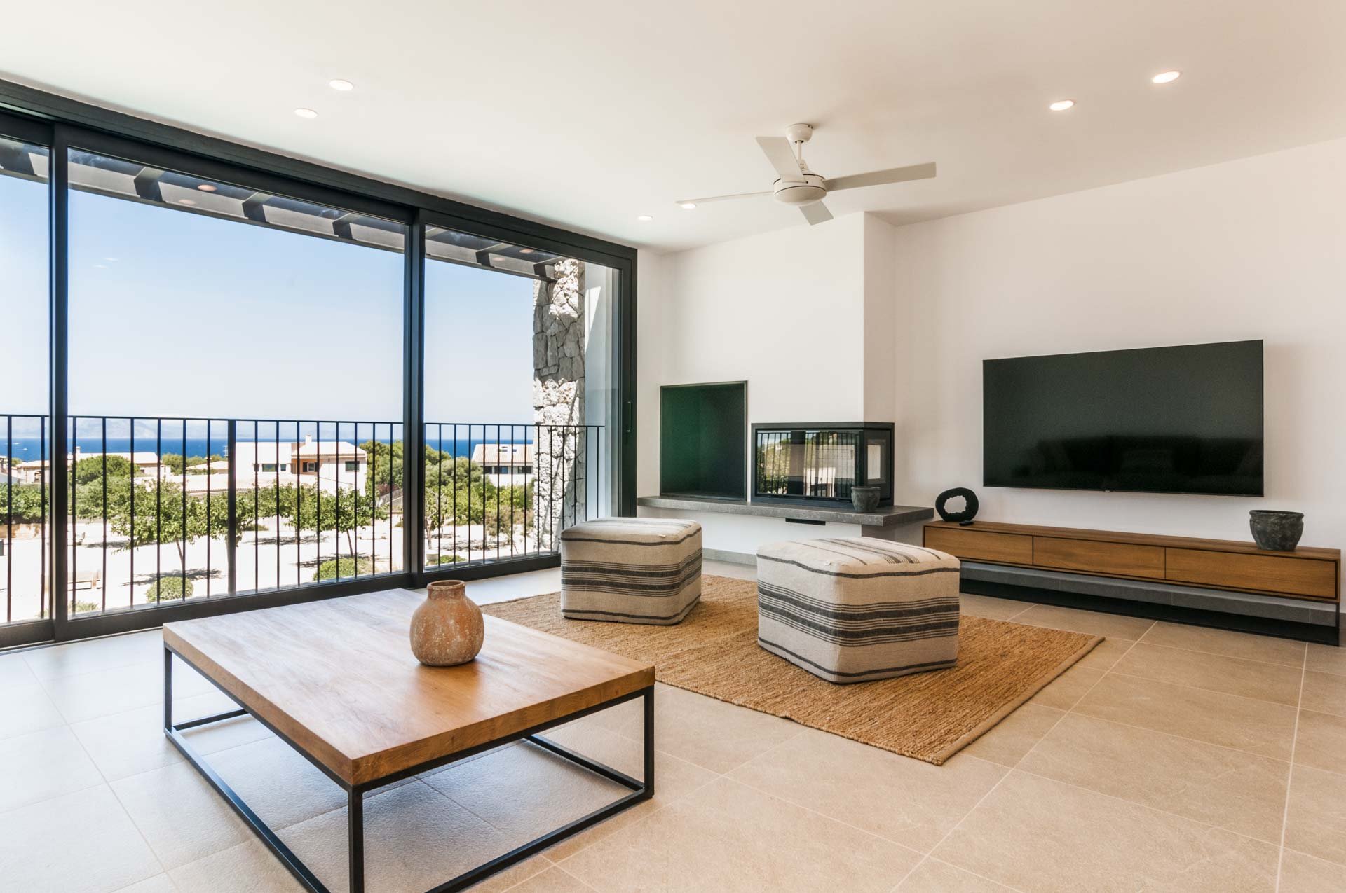 Modern living room with large glass windows overlooking outdoor scenery, featuring a ceiling fan, flat-screen TV, two striped poufs, a wooden coffee table, and a fireplace.