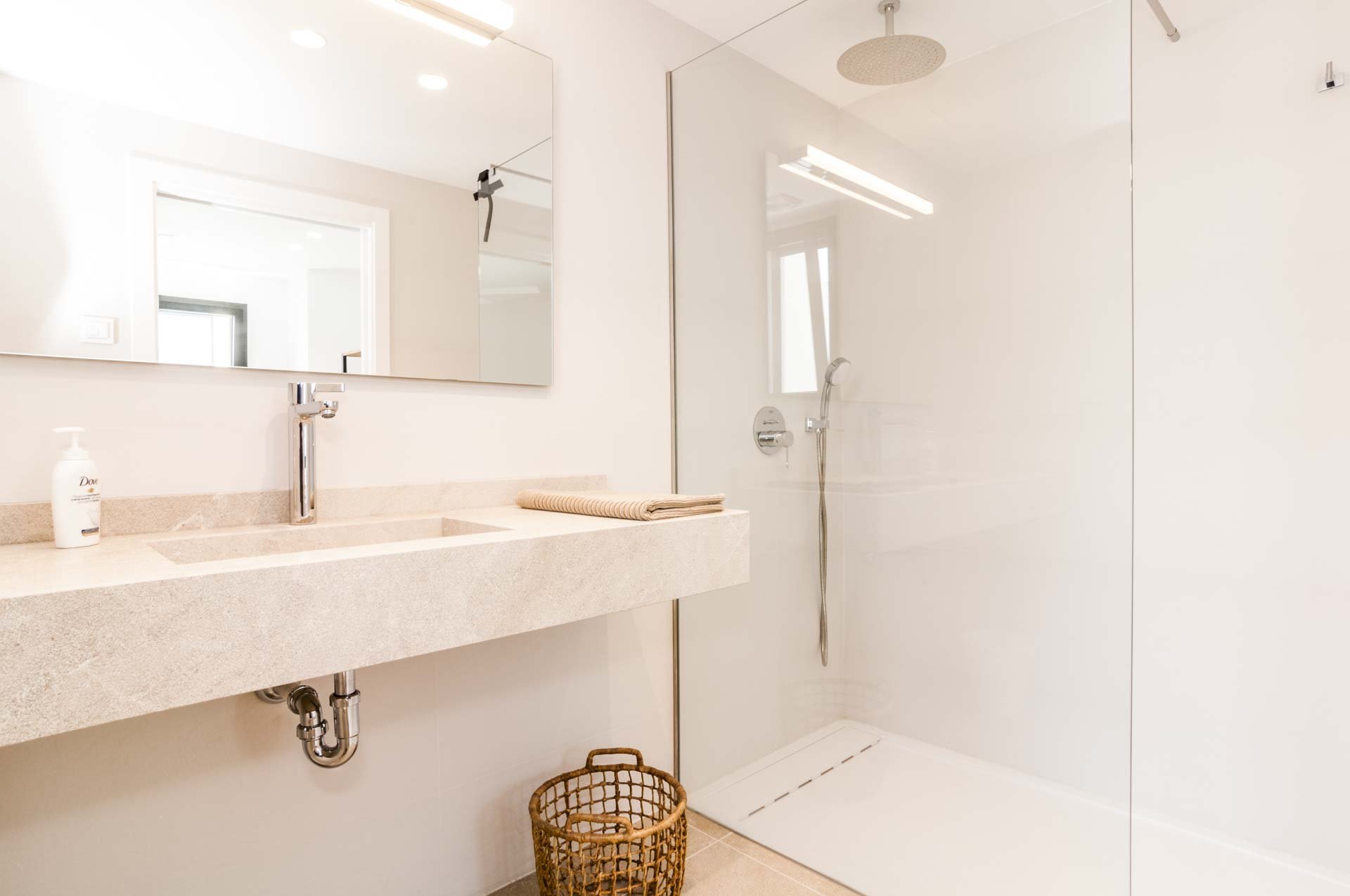 Modern bathroom with beige stone countertop, large mirror, walk-in glass shower, and wicker basket.
