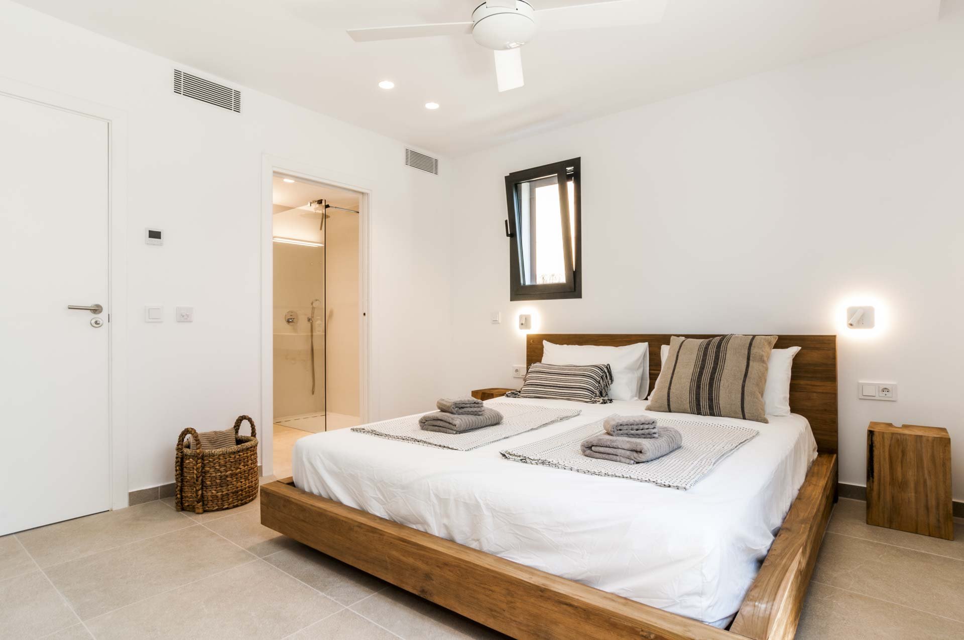 Modern bedroom with a wooden bed, neatly folded gray towels, striped pillows, and an ensuite bathroom with glass door, featuring a white ceiling fan and basket near the door.