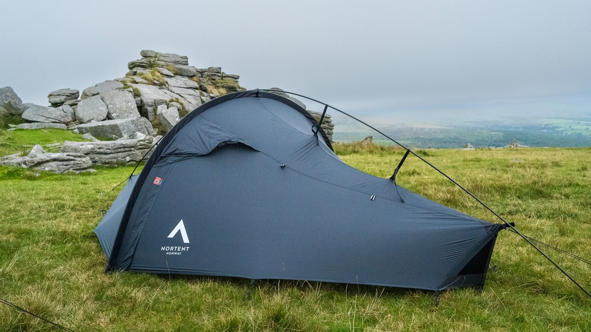 A dark gray tent set up outdoors in a grassy area with rocks and a foggy, mountainous landscape in the background.