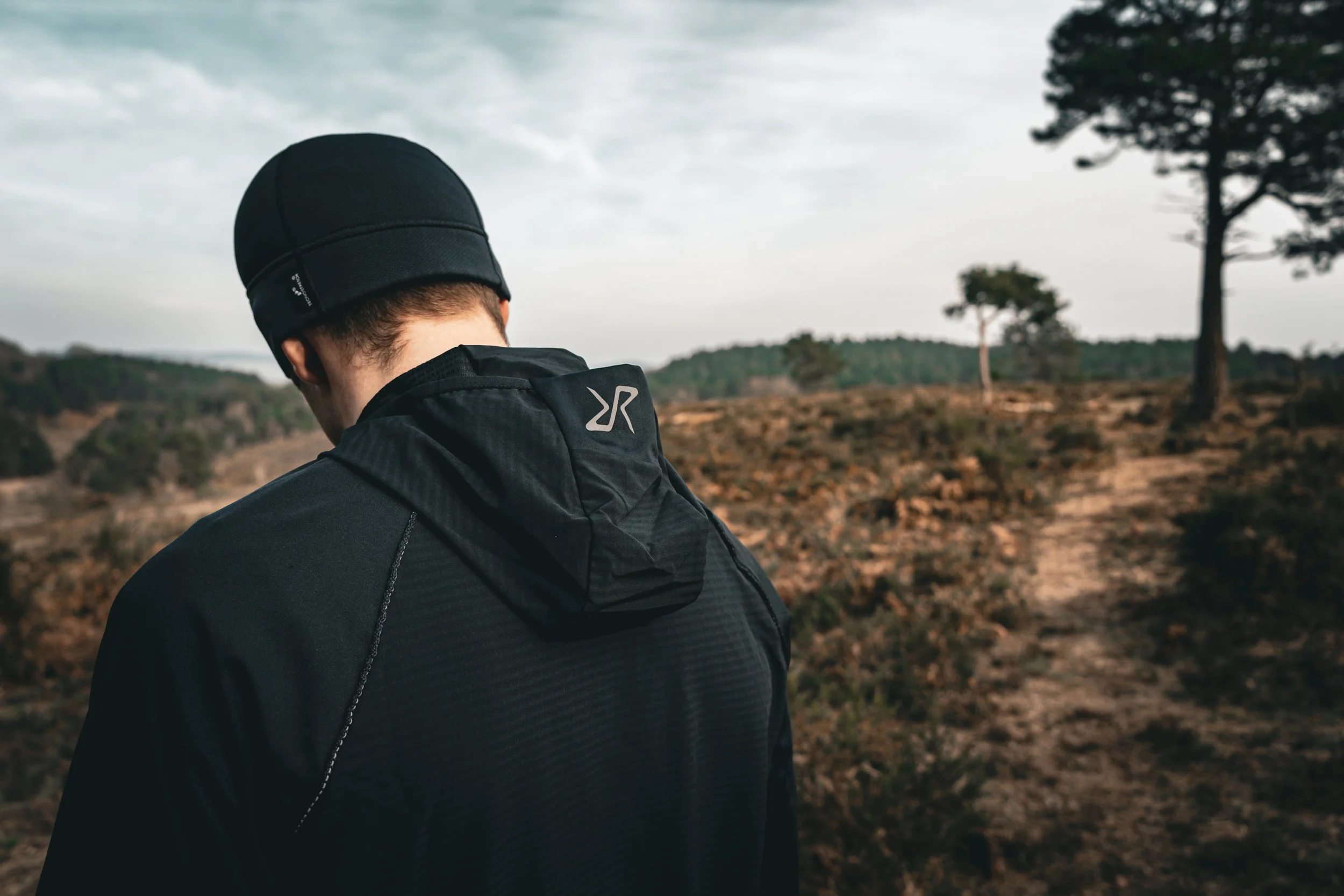 A person wearing a black beanie and black jacket with a logo on the back, standing outdoors with trees and a cloudy sky in the background.