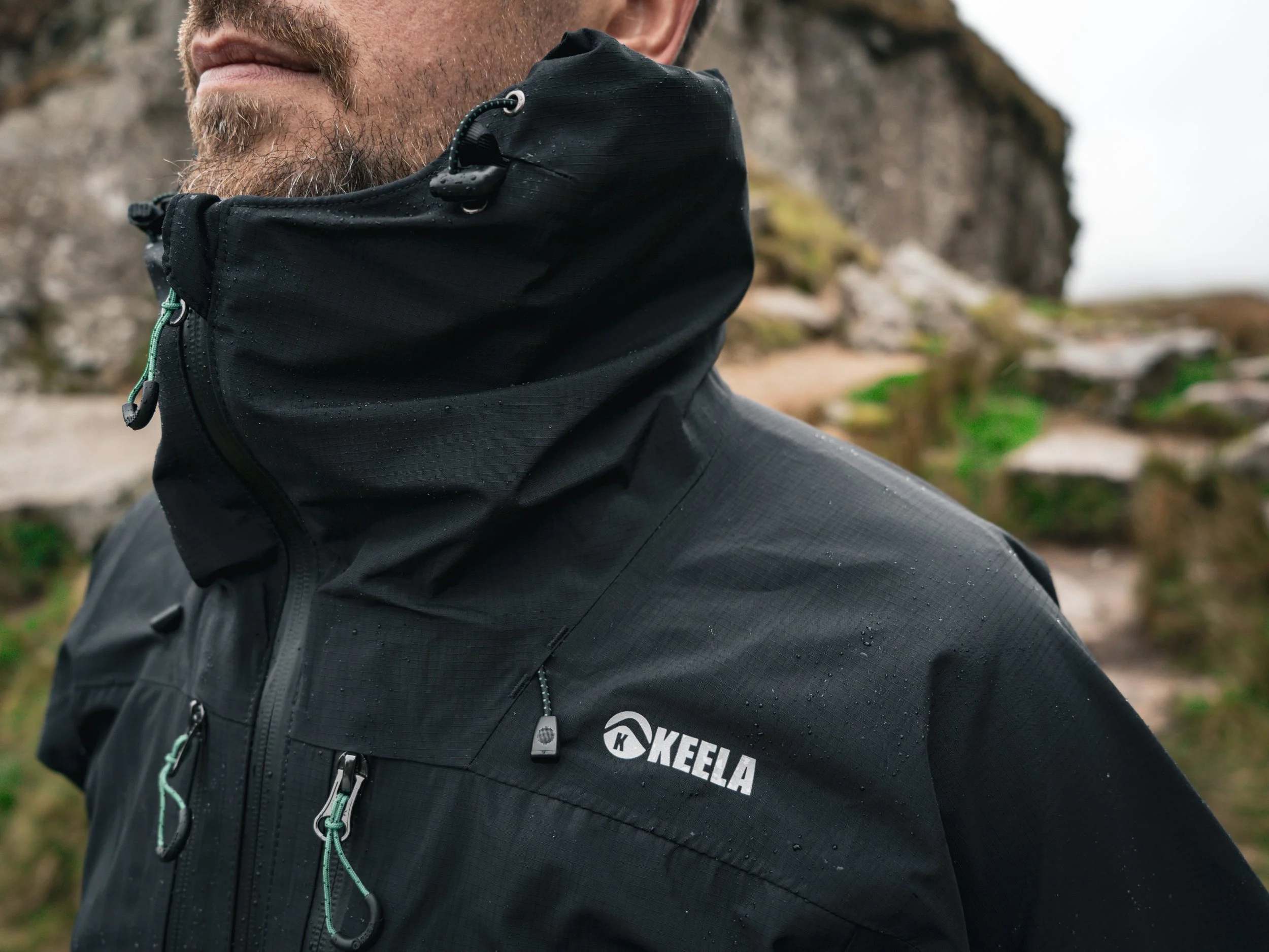 Close-up of a person's face and upper body wearing a black outdoor jacket with a hood, outdoors on a rainy day with wet hair and water droplets visible on the jacket, in a natural setting with rocks and blurry background.
