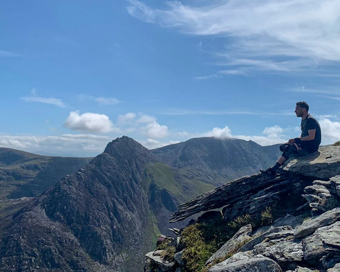 A man sitting on the edge of a rocky outcrop overlooking mountain peaks and a deep valley under a blue sky with clouds.