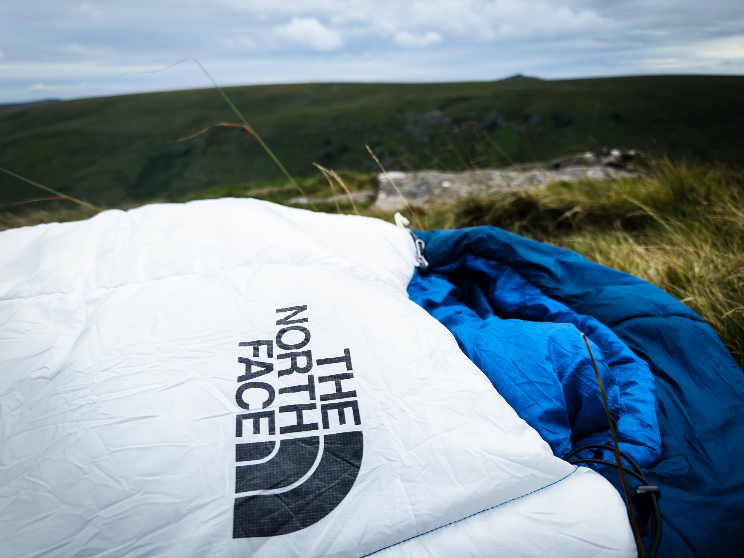 A partially inflated white and blue parachute with the text 'North Face' on it, lying on grassy terrain with hills and a cloudy sky in the background.