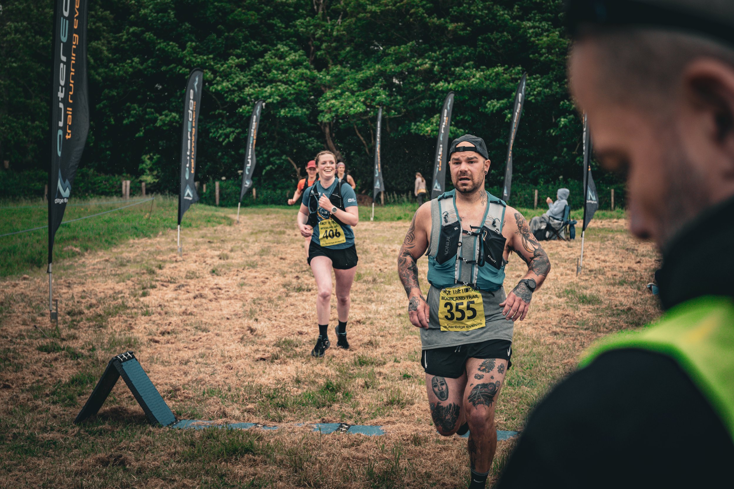 A group of runners participating in an outdoor trail race, with one man in the foreground showing tattoos and a race bib, and a woman smiling as she approaches the finish line, surrounded by trees and race banners.
