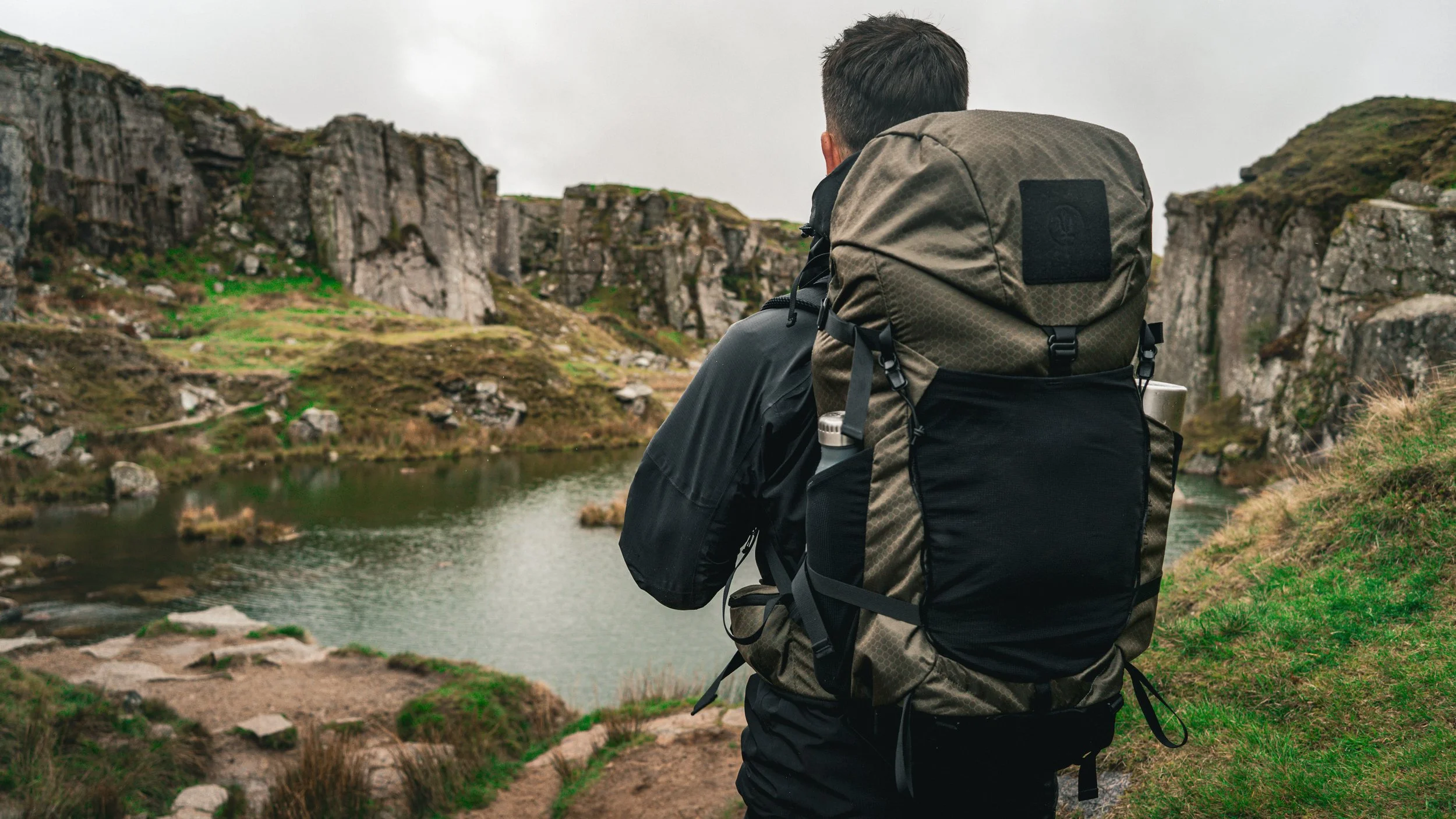 A person with a backpack sitting by a river in a mountainous landscape, viewed from behind.