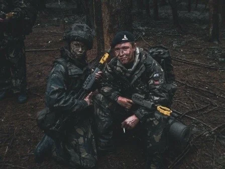 Two soldiers in camouflage gear with face paint holding rifles in a wooded area during daytime.