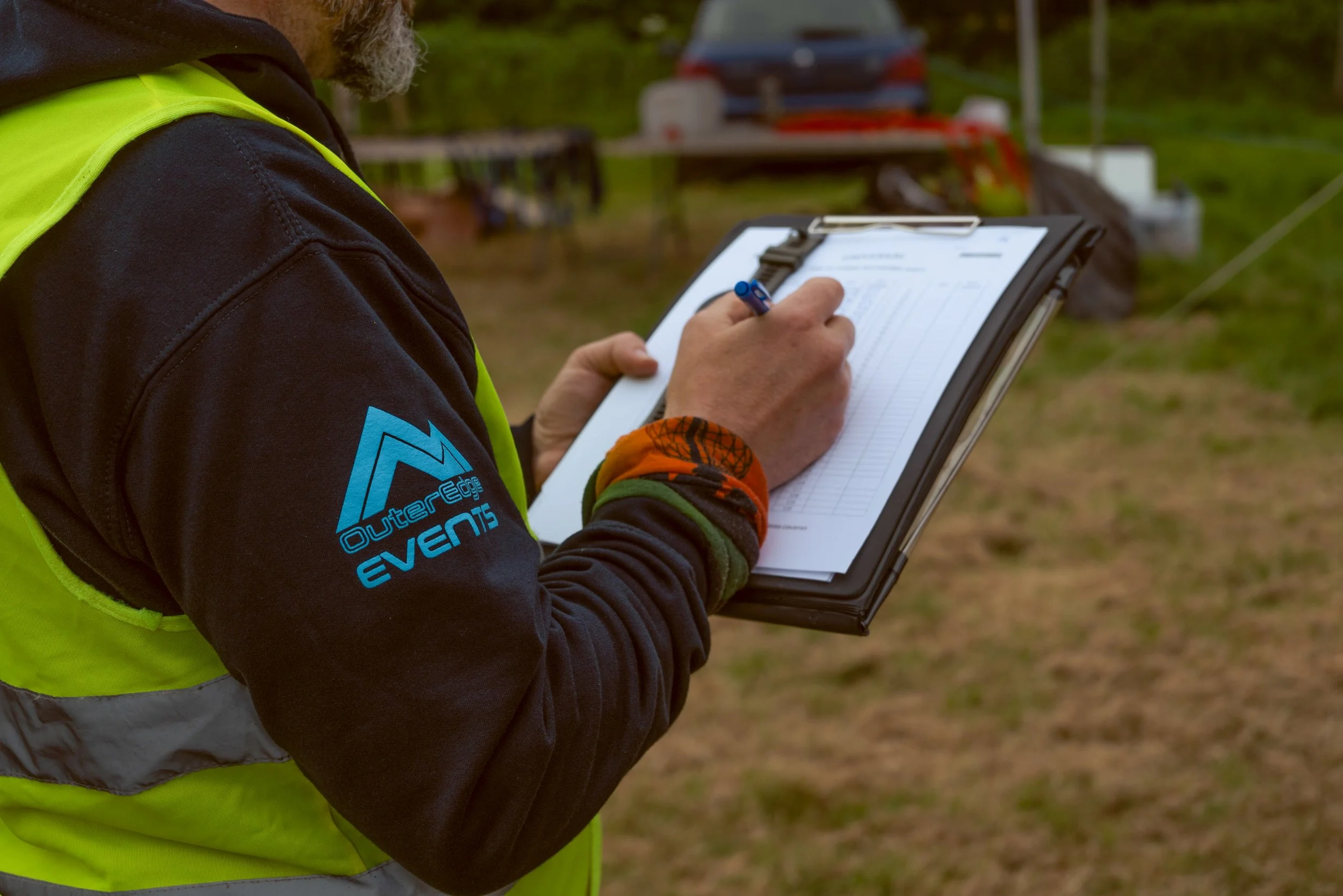 Person in a neon safety vest and black hoodie holding a clipboard and writing with a blue pen outdoors.