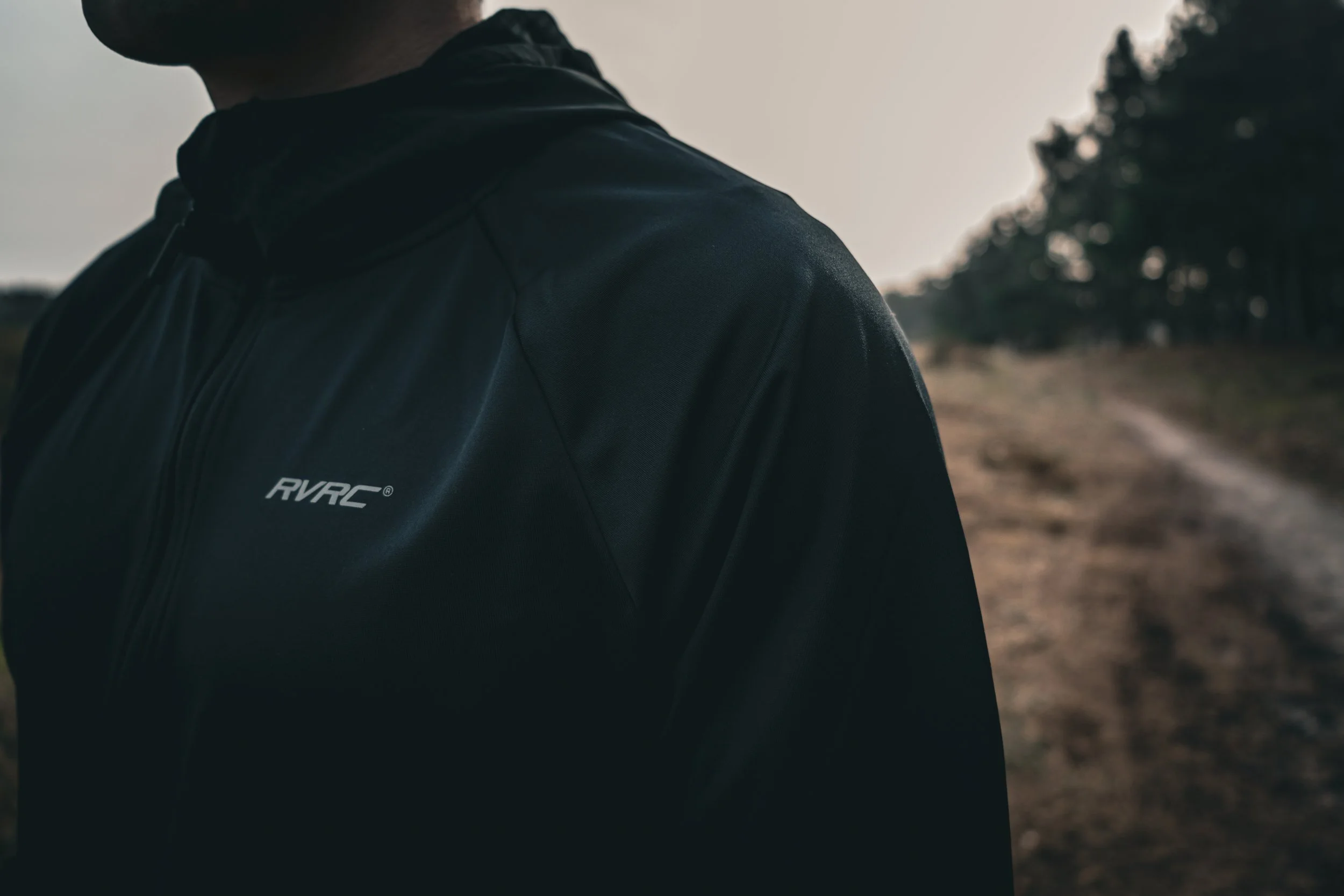 Close-up of a person wearing a black jacket with a logo on it, standing outdoors near trees and a dirt path.