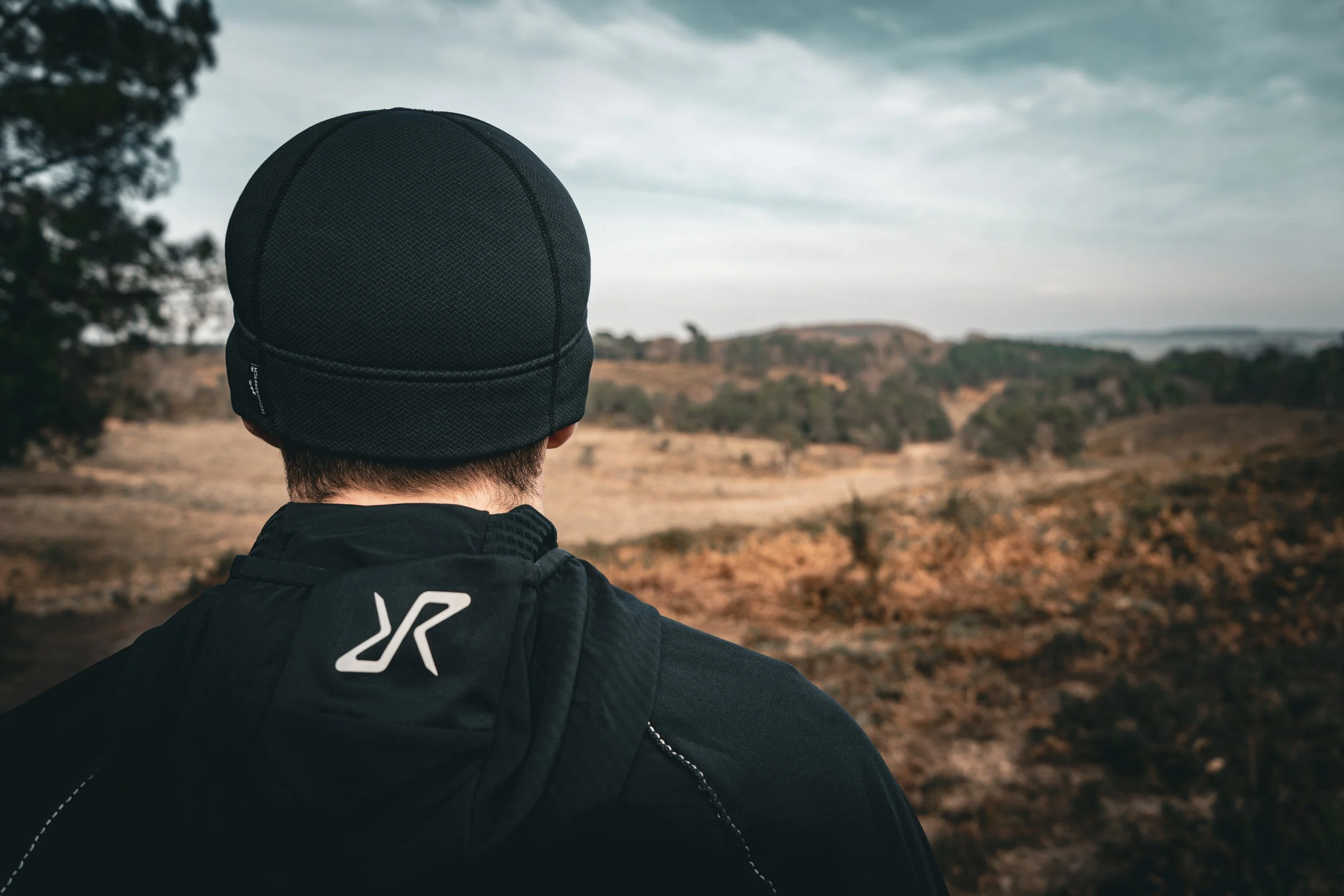 A person wearing a black beanie and black jacket, seen from behind, looking out over a hilly landscape with sparse trees and cloudy sky.