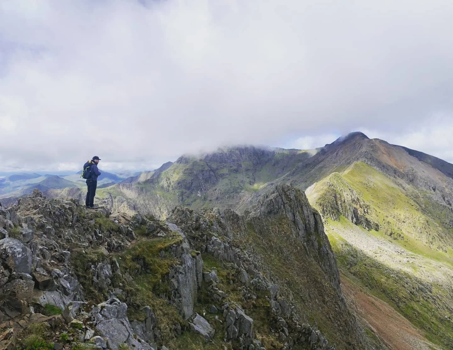 A person standing on rocky mountain terrain overlooking green mountains and cloudy sky.