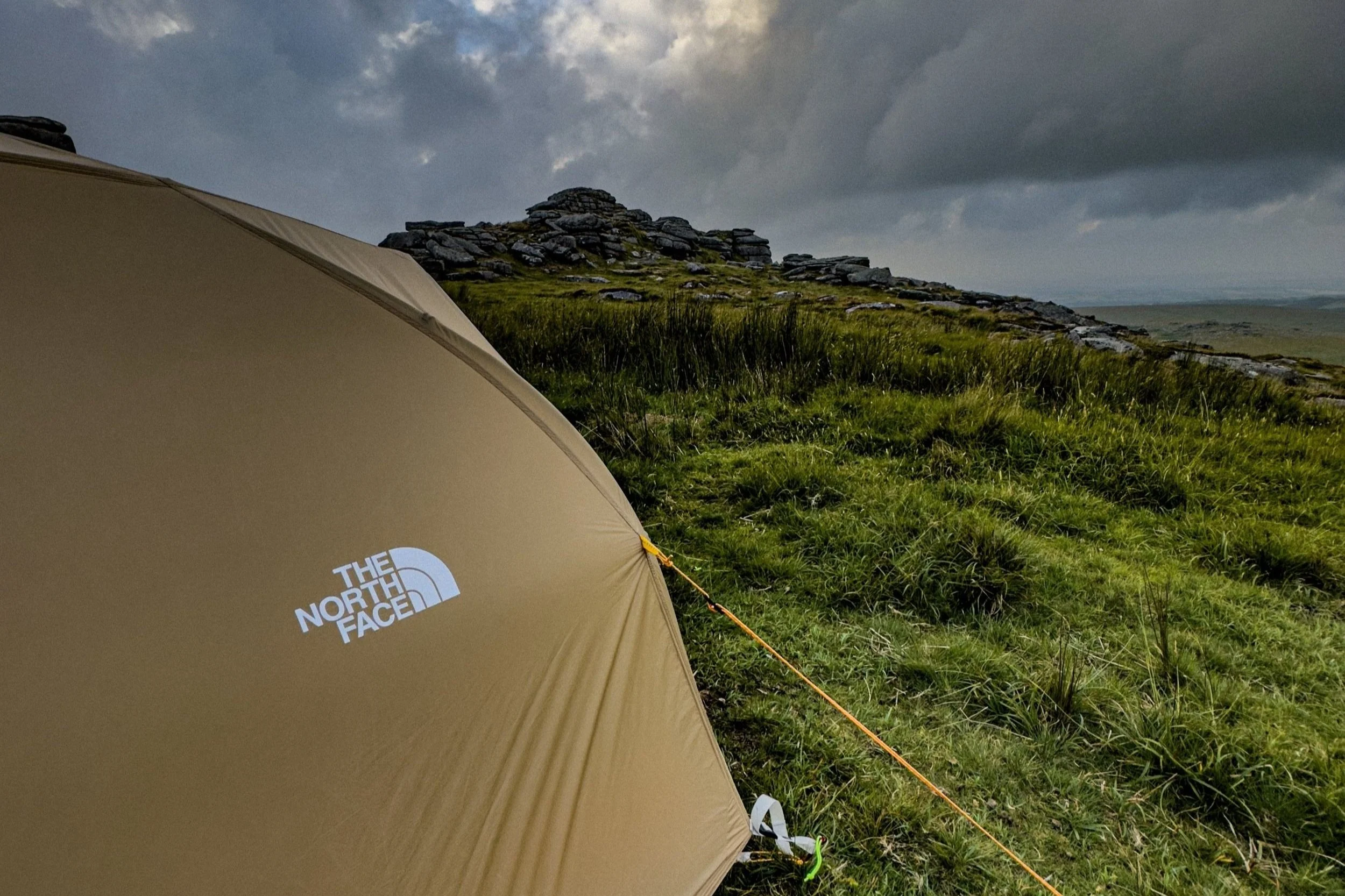 A beige North Face tent set up on lush green grass with a rocky hill and cloudy sky in the background.