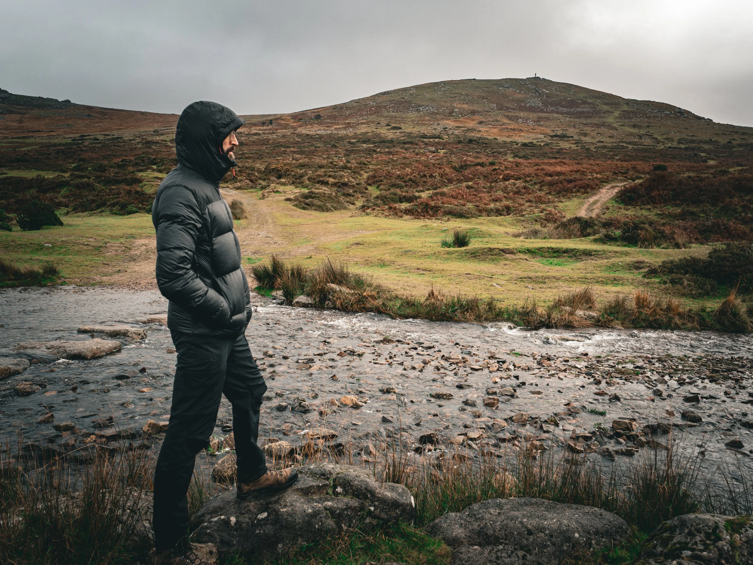 A man wearing a black hooded jacket, khaki pants, and hiking boots standing on rocks near a stream in a hilly landscape during overcast weather.