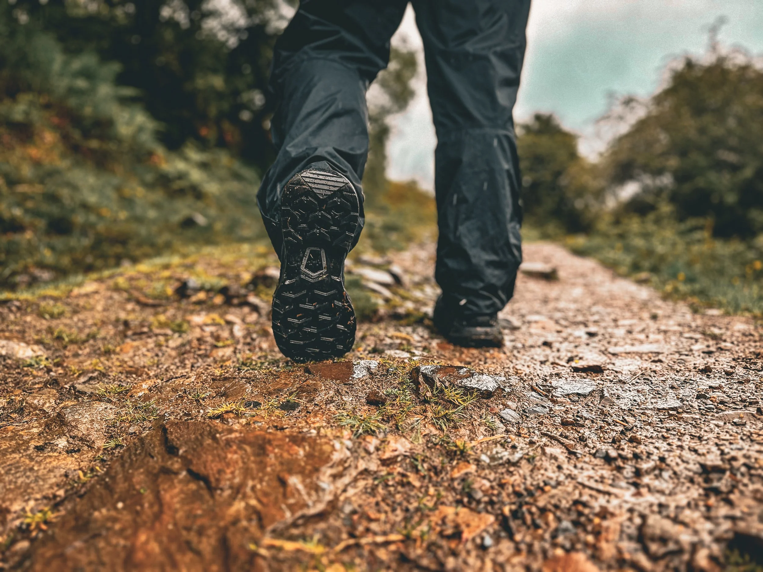 Close-up of a hiker's feet in rugged hiking boots walking on a dirt trail in a forested area with trees and autumn leaves.