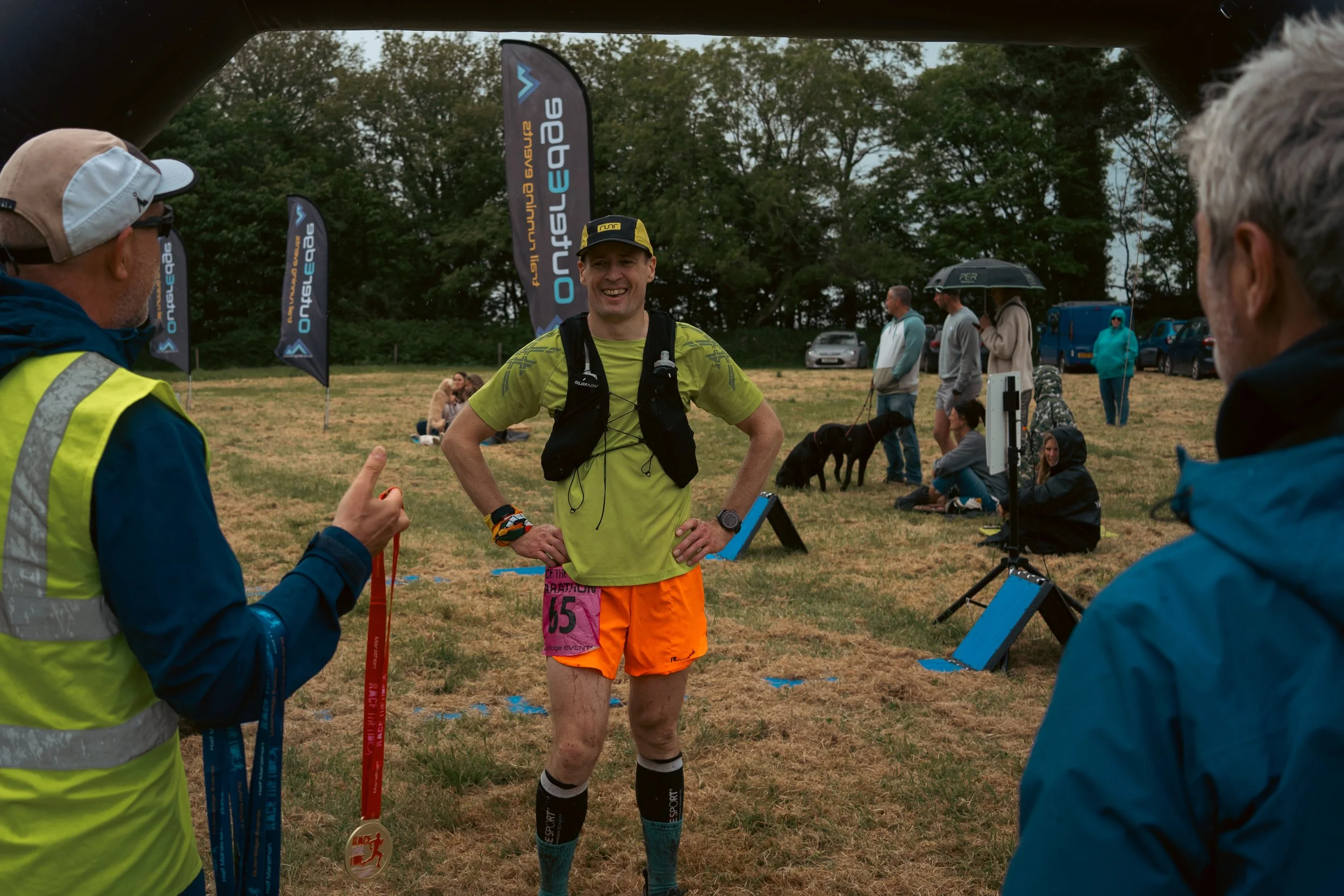 A smiling male runner in bright running gear standing after finishing a race, being interviewed by a person holding medals, with race flags and other spectators in the background at an outdoor event.