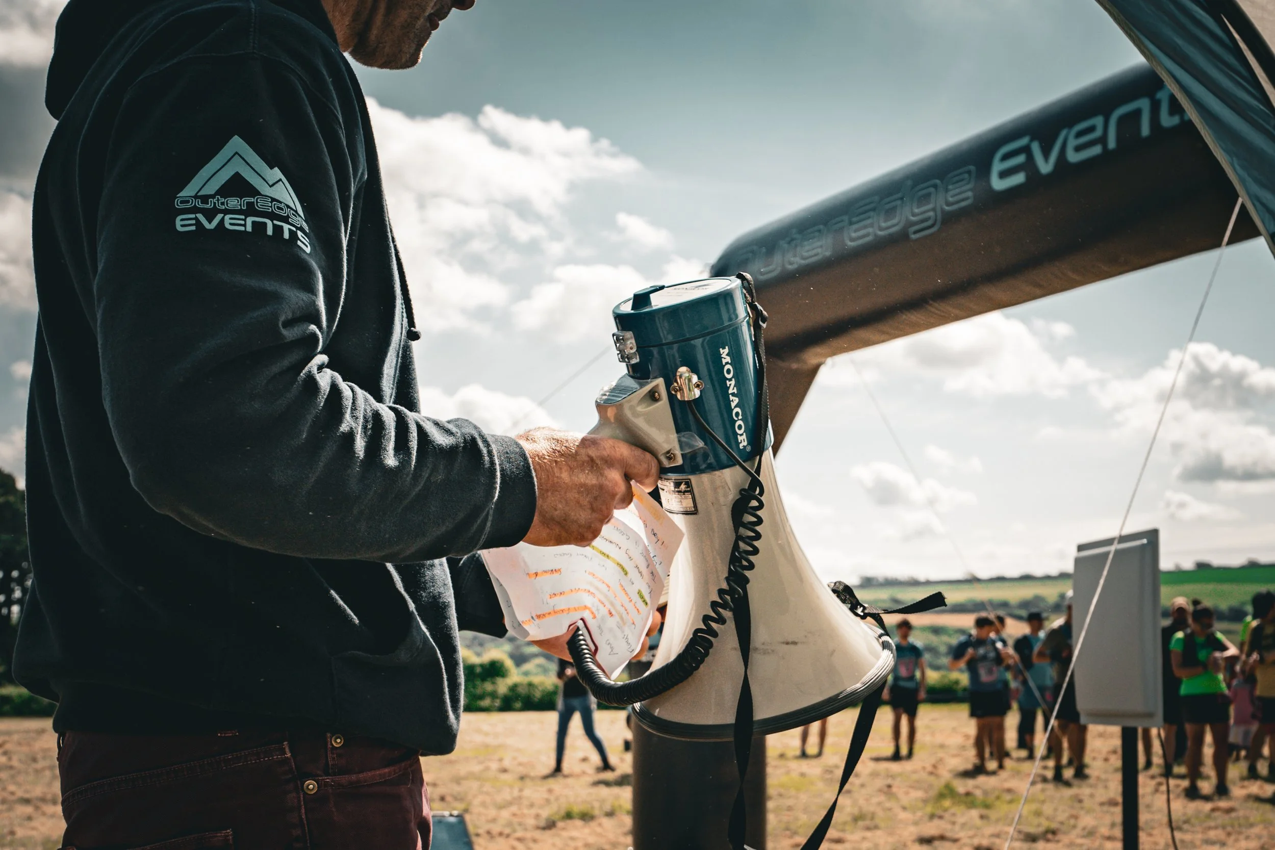A man holding a document stands next to a person operating a drone outdoors, with a group of people in the background and a partially visible drone or aircraft.