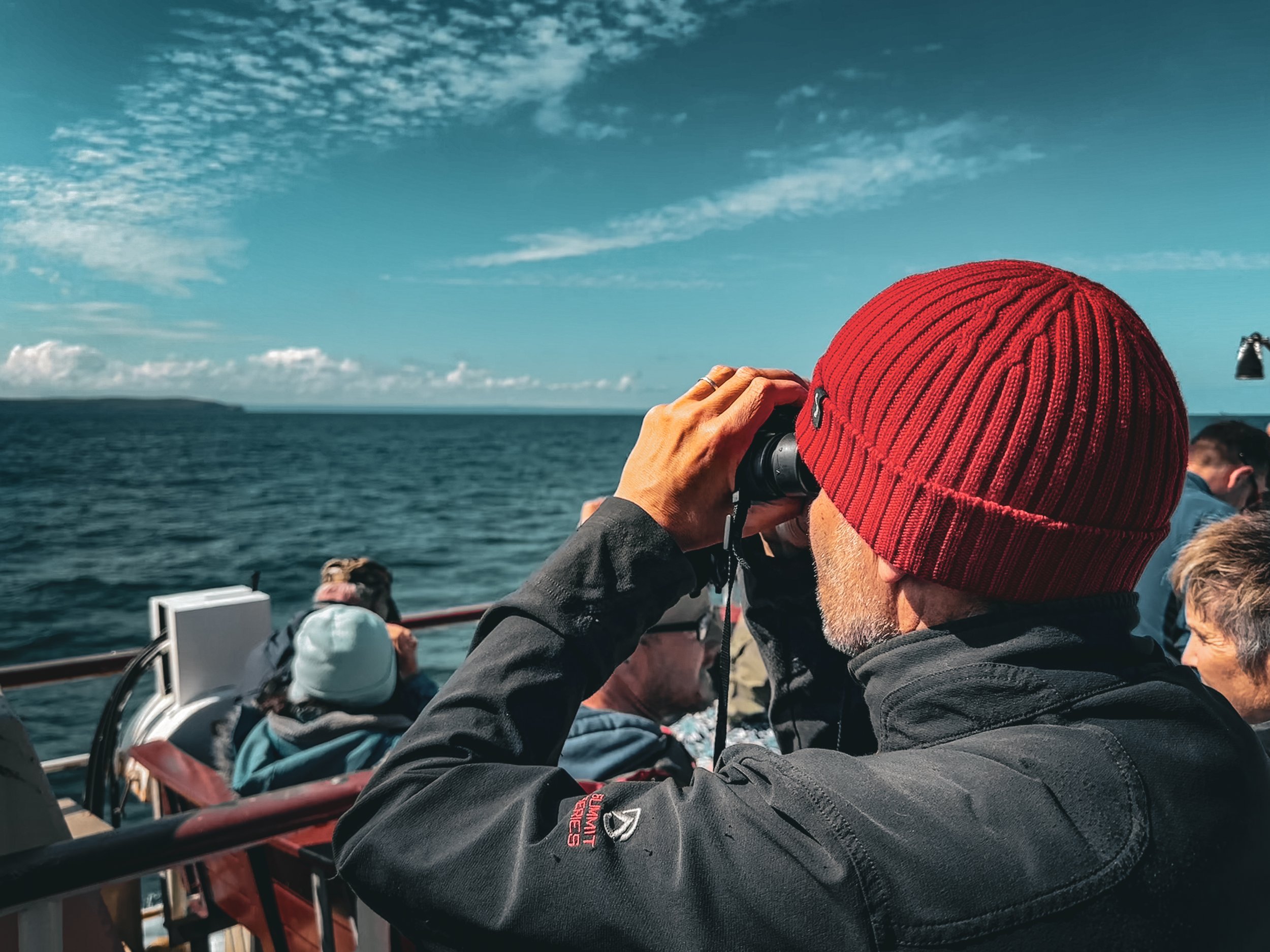 People on a boat in the water, with one person holding binoculars, wearing a red knit hat, and others sitting or standing around, under a partly cloudy sky.