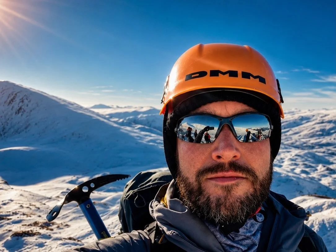 A man with a beard wearing a helmet, sunglasses, and winter gear, standing in snowy mountains during a sunny day, with their reflection visible in the sunglasses.