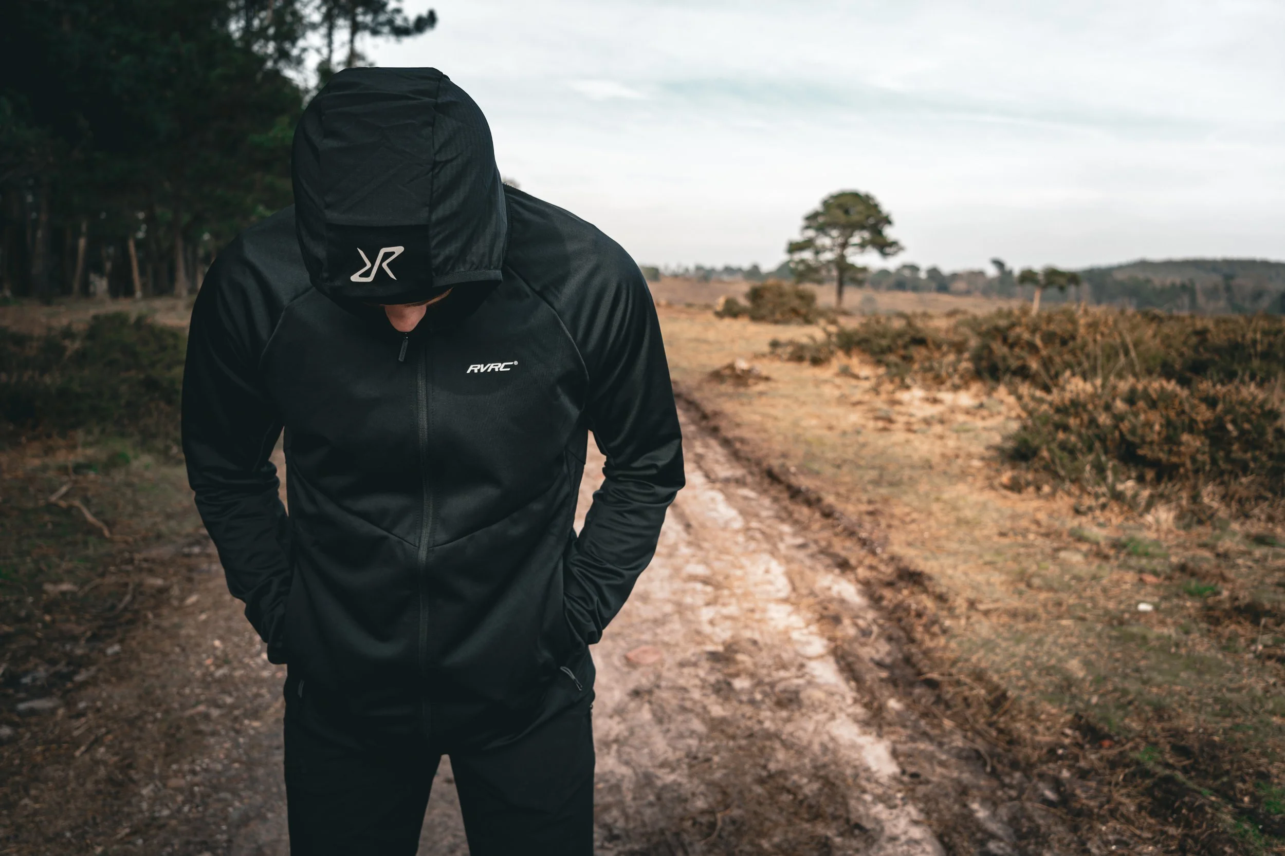 Person dressed in black athletic wear with a hoodie pulled over their head, walking on a dirt trail in a rural landscape with sparse vegetation and trees in the background.