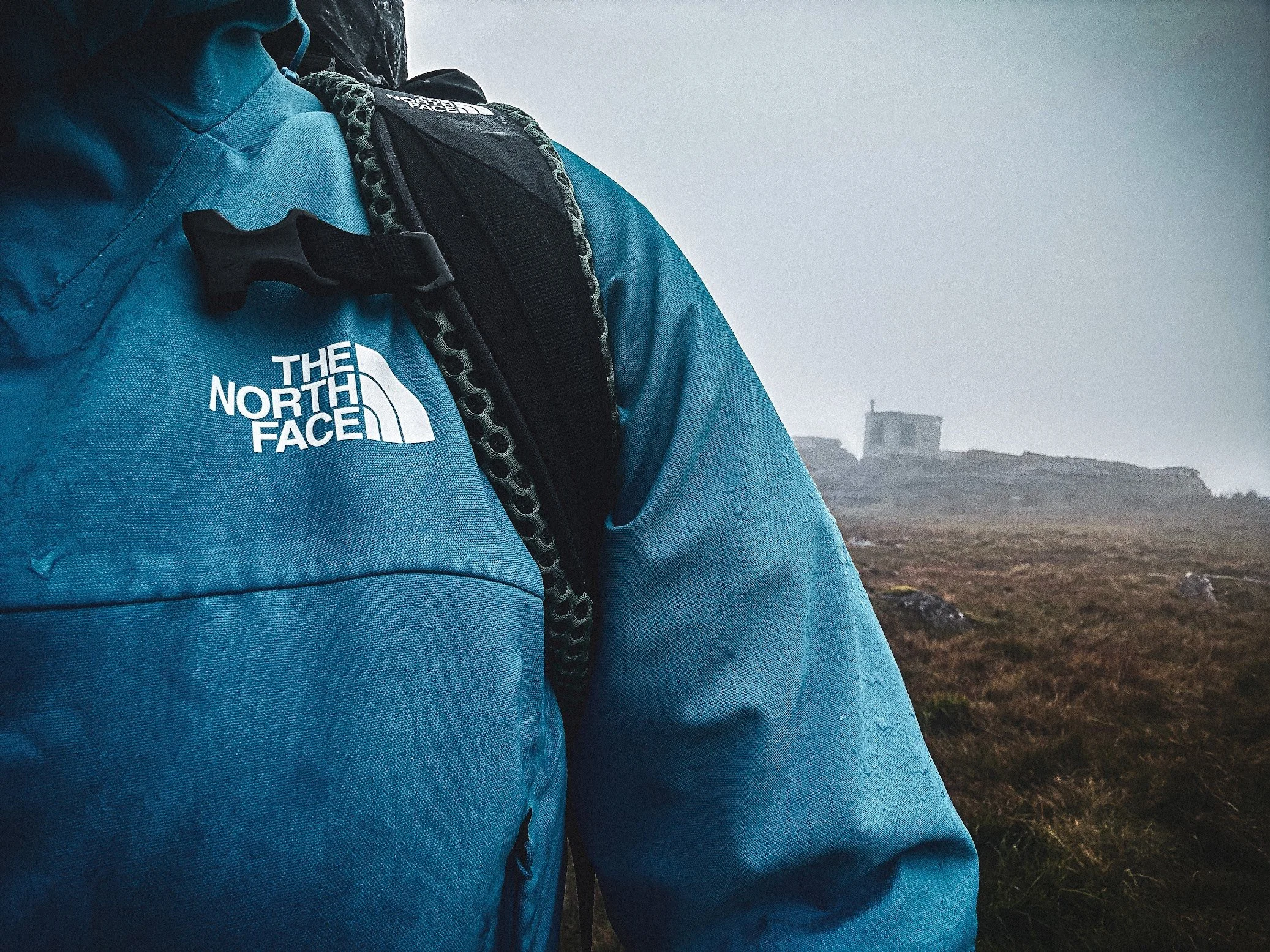 Close-up of a person wearing a teal North Face jacket and a black backpack, standing outdoors in a foggy landscape with a building in the distance.