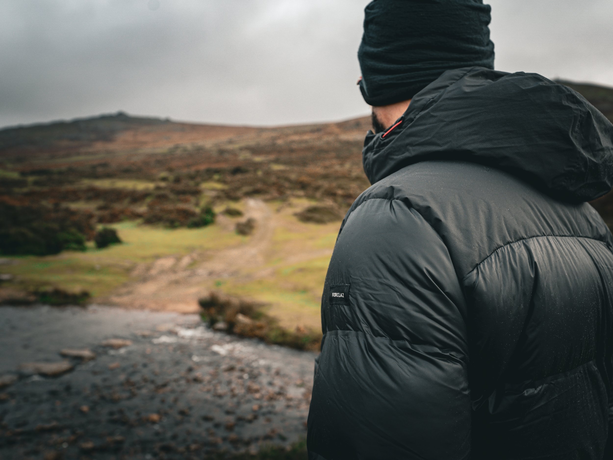 A person dressed in a black jacket and black beanie looking out over a rugged landscape with hills and a cloudy sky, possibly on a hike.