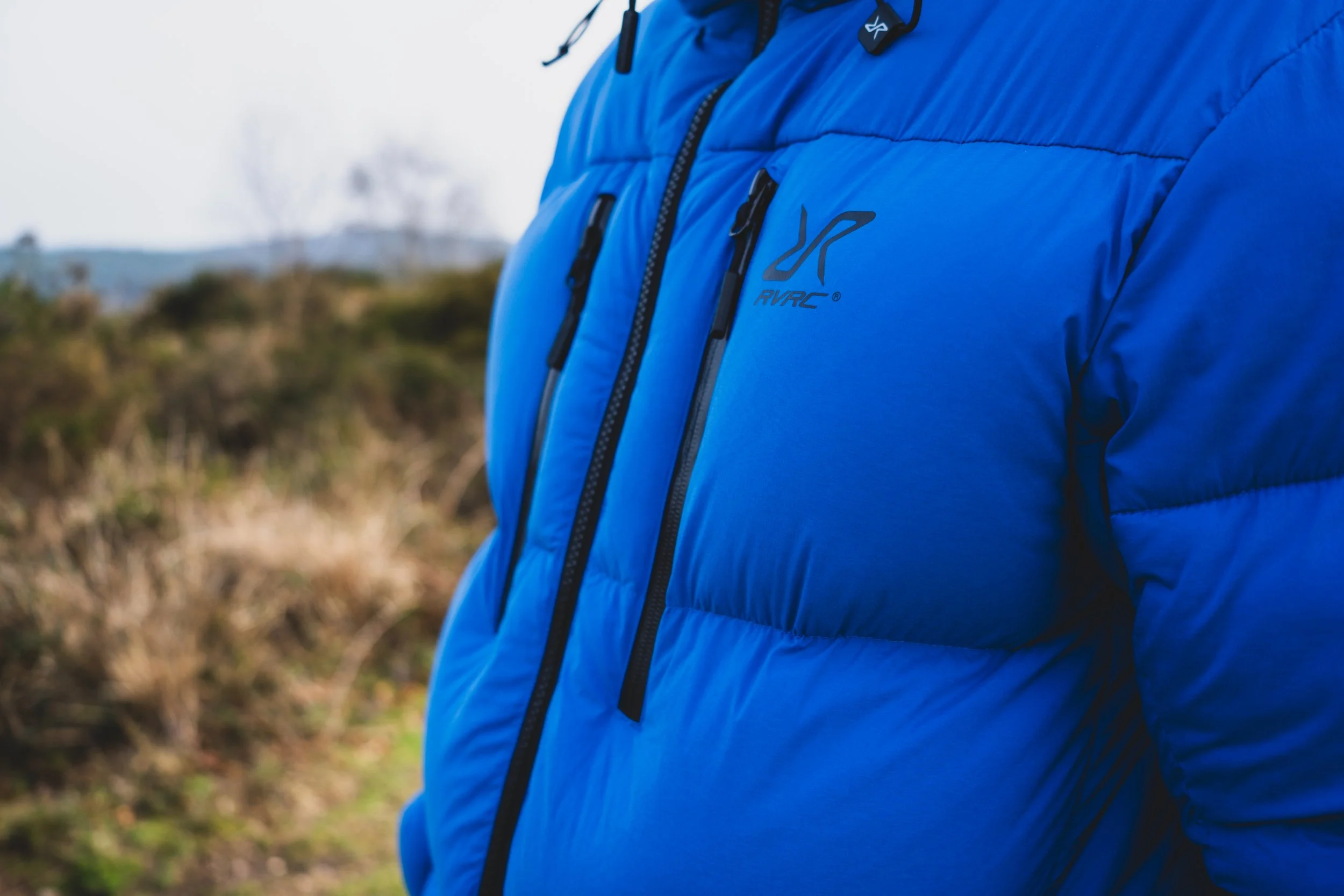 Close-up of a person wearing a bright blue puffer jacket with black zippers outdoors in a natural, blurred background.