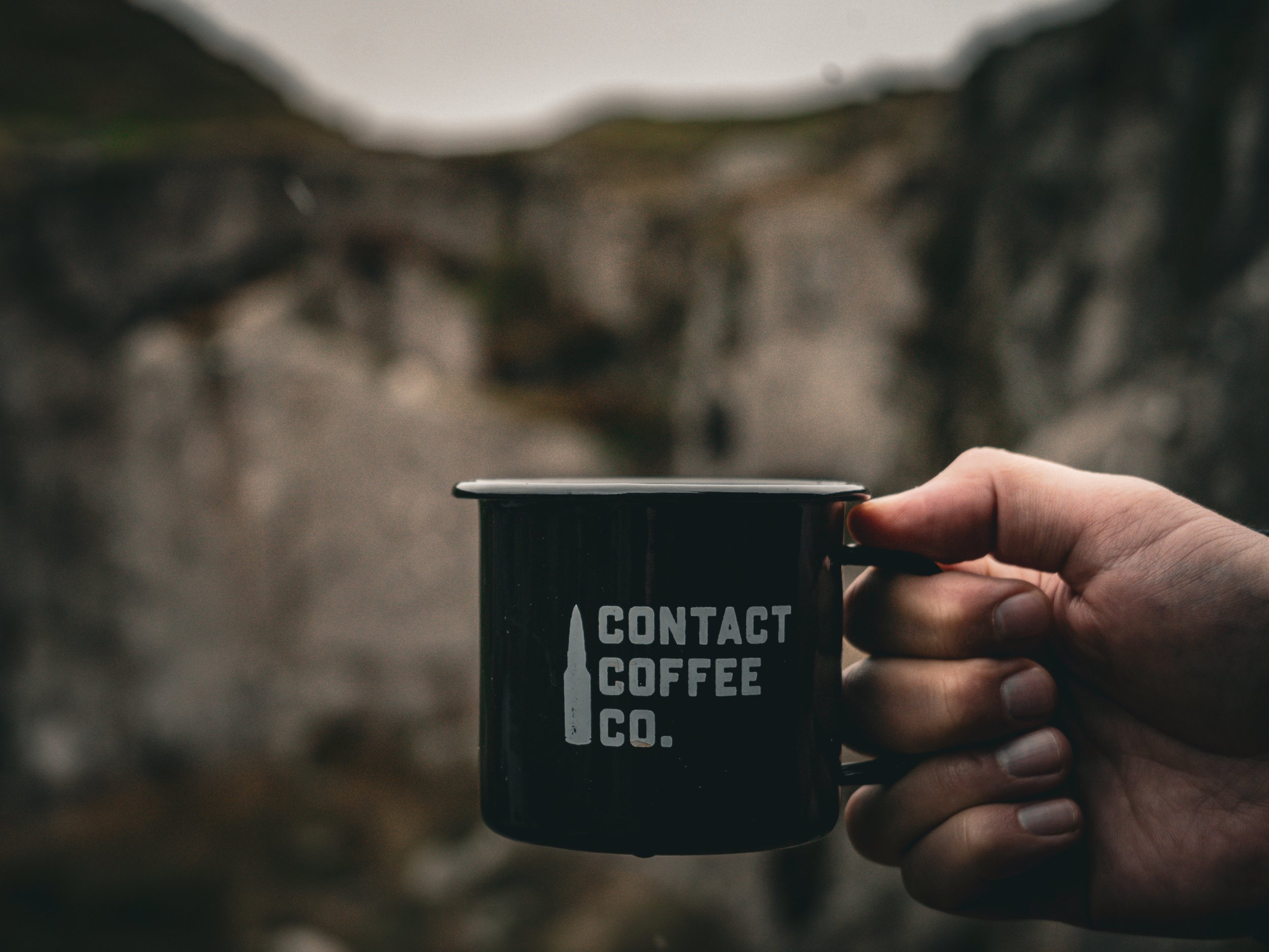 A person holding a black coffee cup with white text that says "Contact Coffee Co." outdoors with a blurred natural background.