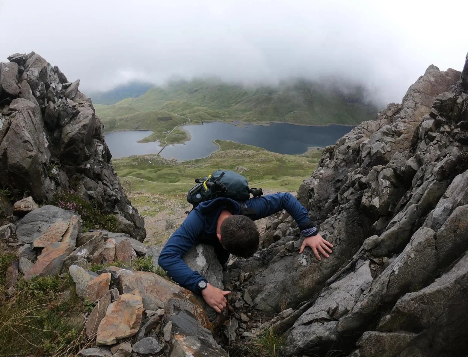 A person climbing a rocky mountain with a backpack, overlooking a scenic view of lakes and green hills, with clouds partly covering the sky.